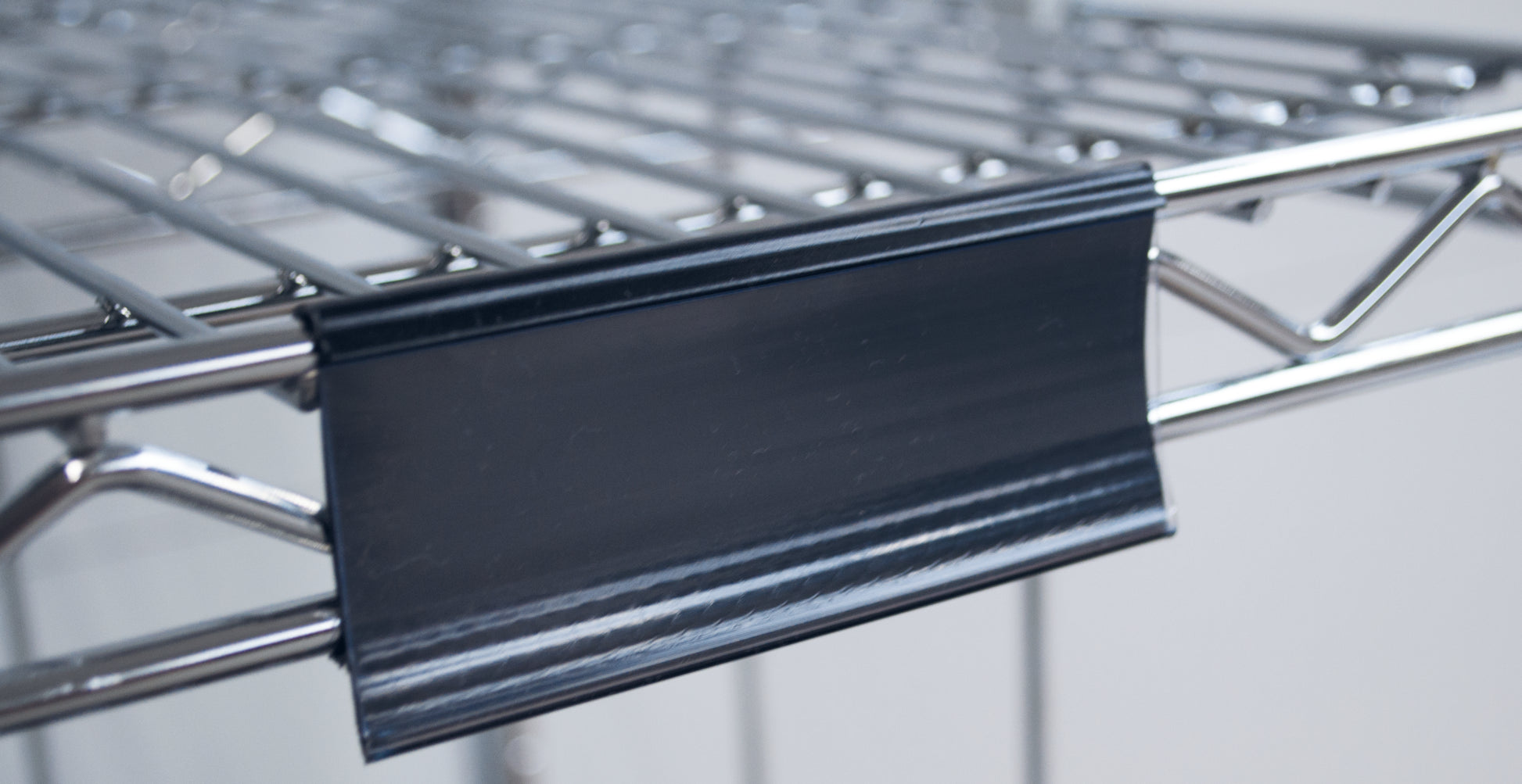 Nahaufnahme eines Novocal Etikettenrahmens mit Papierschildern (ca. 100 x 44 mm, 1 Stück), der an einem Metalldrahtregal befestigt ist, mit dem schwarzen Kunststoffetikettenhalter an der Vorderkante; der Hintergrund ist unscharf.