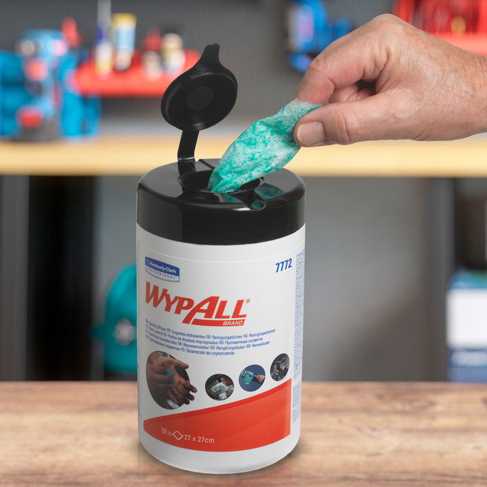 Eine Hand zieht ein grünes WypAll® Wischtuch mit hoher Saugfähigkeit aus einer offenen Wanne der Kimberly-Clark GmbH (6 Wannen x 50 Blatt). Im unscharfen Hintergrund sind Regale mit verschiedenen Artikeln zu sehen.