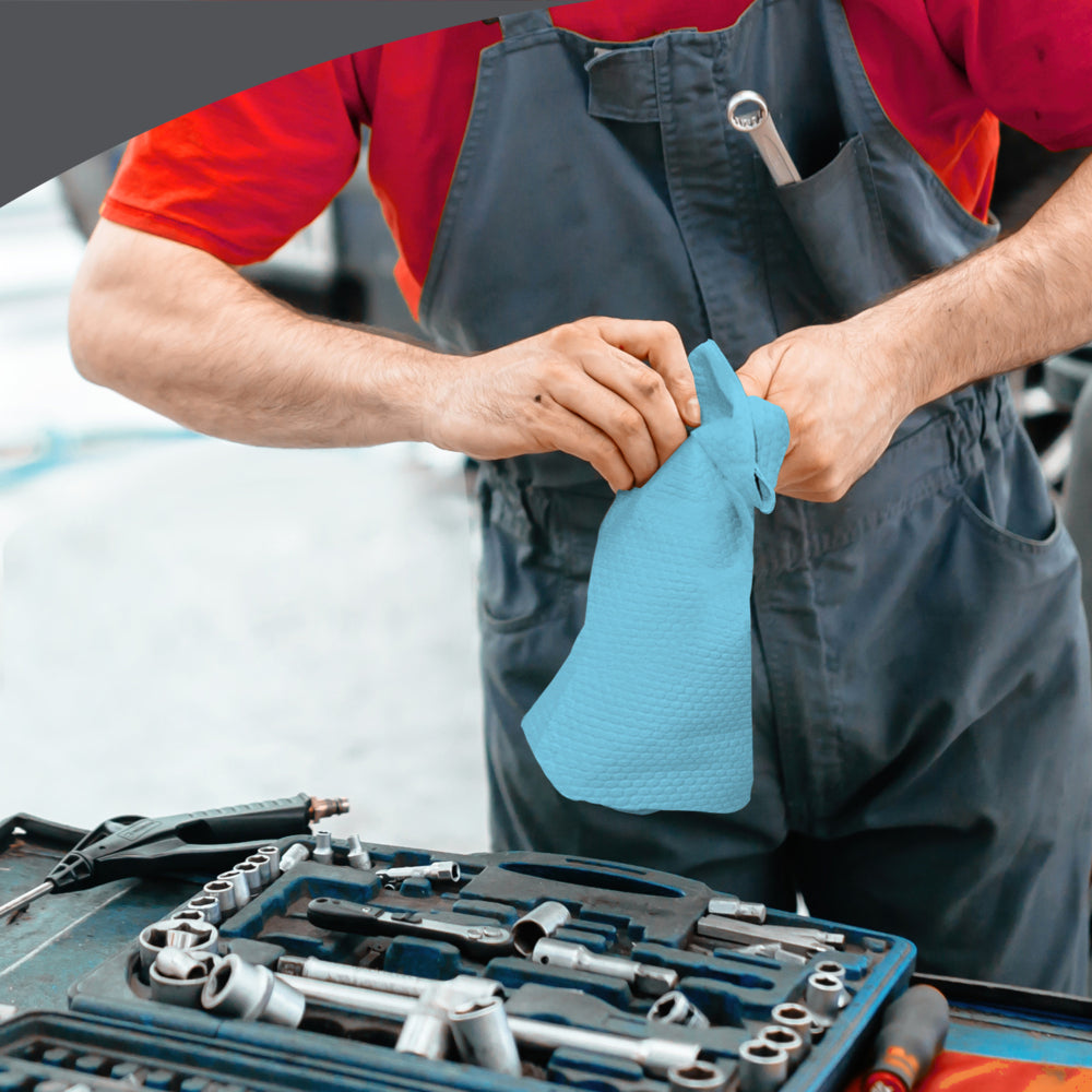 Eine Person in grauem Overall und rotem Hemd wischt sich mit einer WypAll® X70 PowerClean - Zupfbox (Blau) der Kimberly-Clark GmbH die Hände neben einer mit Werkzeugen bestückten Werkbank ab und sorgt so für professionelle Reinigung am Arbeitsplatz.