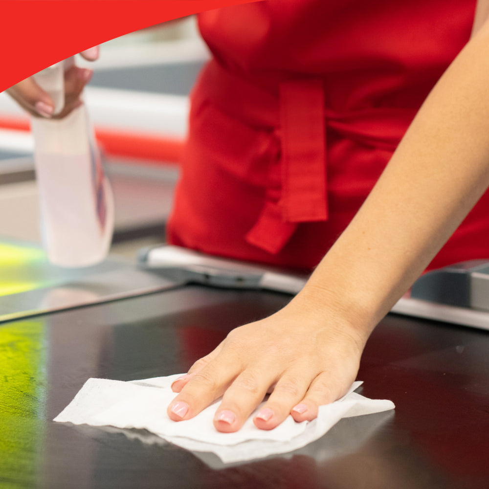 Eine Person mit roter Schürze reinigt eine Arbeitsplatte mit einem Tuch aus der WypAll® X50 General Clean Großrolle (1 Rolle x 1100 Blatt) der Kimberly-Clark GmbH und hält eine Sprühflasche für die effiziente Oberflächenreinigung in der Hand.