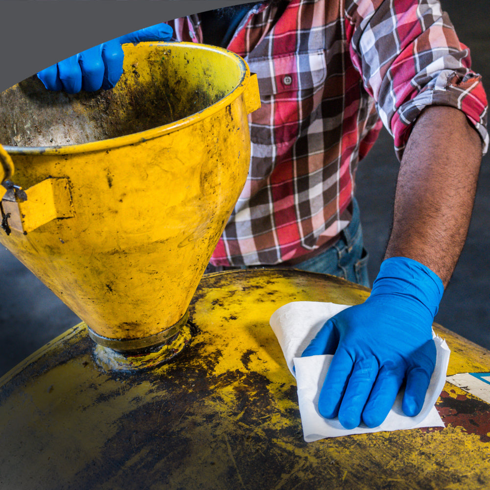 Eine Person mit blauen Handschuhen und kariertem Hemd wischt einen gelben Industriecontainer mit einem Kimberly-Clark WypAll® X70 PowerClean Tuch (Weiß, 1 Box x 300 Blatt) neben einem gelben Trichter für gründliche industrielle Reinigung.