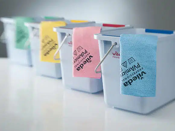 Vier weiße Putzeimer stehen aufgereiht, über deren Rand jeweils ein andersfarbiges Vileda Professional PVAmicro Microfasertuch hängt. Die blauen, rosa, gelben und grünen Tücher der Vileda GmbH zeigen unterschiedliche Markenlogos.