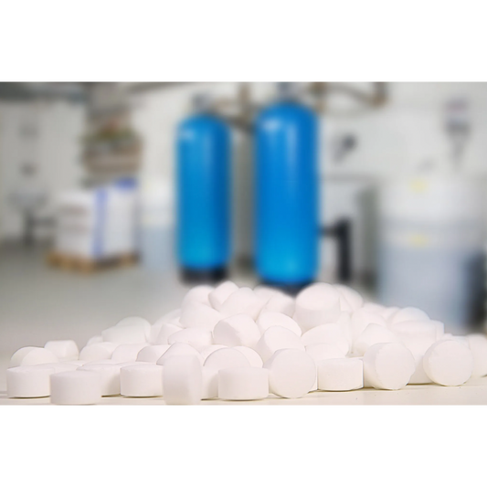 Im Vordergrund fällt ein Stapel Regenit Siedesalztabletten, gemäß EN 973 Typ, der K+S Minerals and Agriculture GmbH auf, während der unscharfe Hintergrund zwei große blaue Wasserenthärtungsanlagen erkennen lässt. Die industrielle Kulisse lässt auf einen präzisen Nutzraum schließen, der den Normen EN 973 Typ A entspricht.