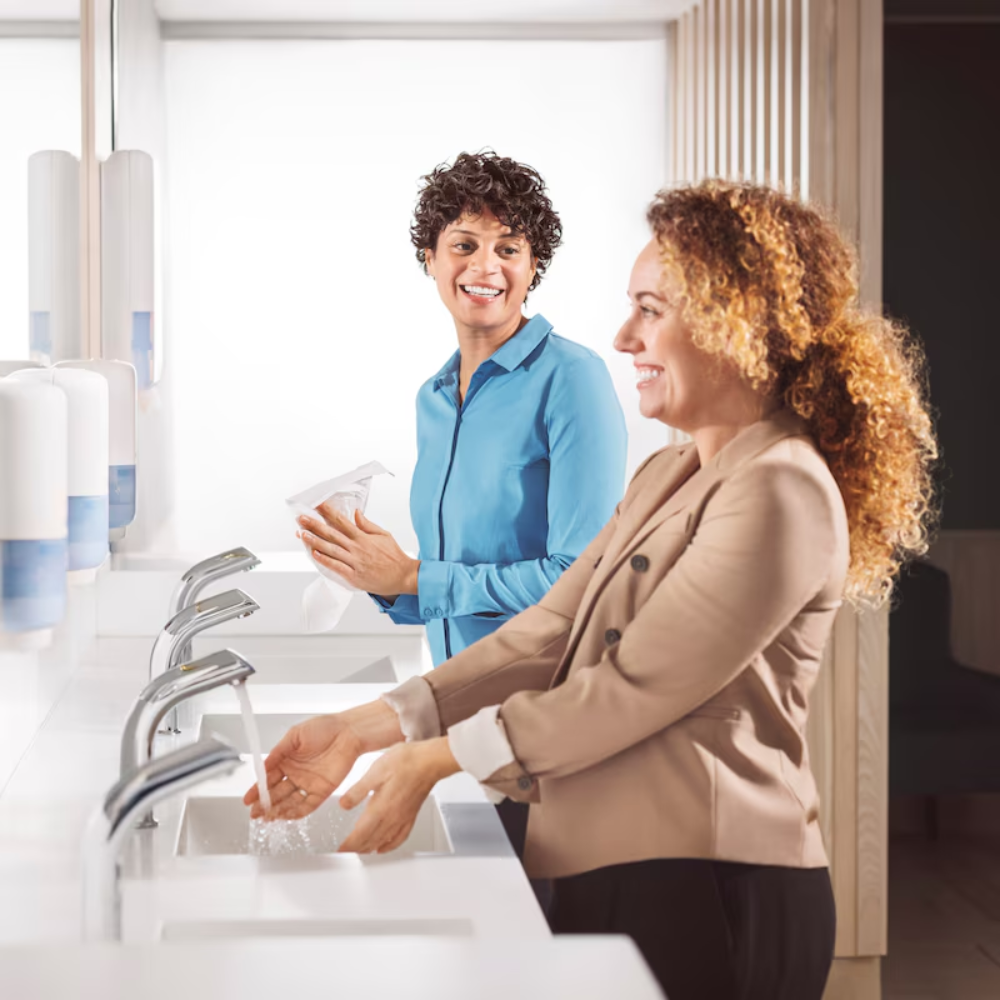 Zwei Frauen stehen an den Waschbecken einer öffentlichen Toilette. Eine wäscht sich die Hände mit Tork 520201 Reine Hand Schaumseife Premium S4 (1000 ml) von TORK – Essity Professional Hygiene Germany GmbH, die andere lächelt und hält Papiertücher in der Hand. Auf dem Waschtisch stehen Seifenspender.