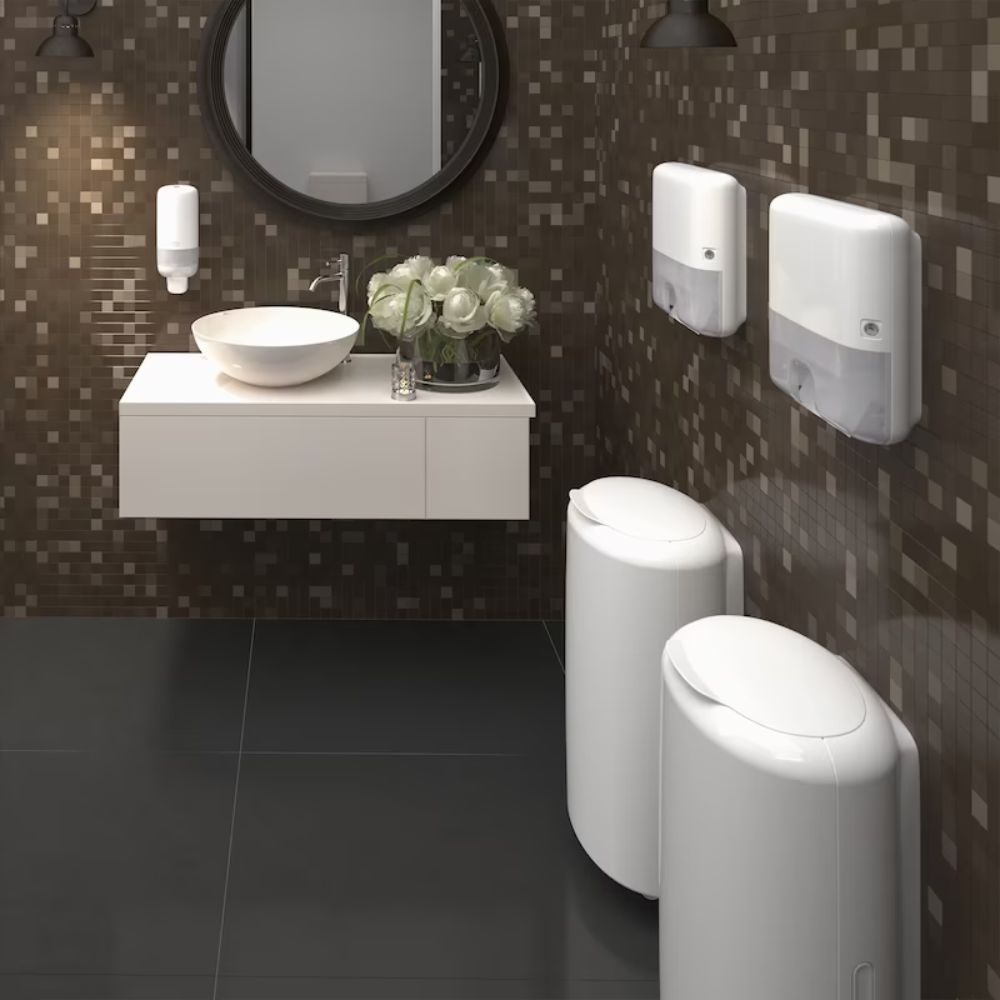 In einem modernen Badezimmer mit einem runden Spiegel über einem weißen, schwebenden Waschtisch und einem Aufsatzwaschbecken sind zwei elegante Tork 561500-Spender für Seife und Handdesinfektionsmittel von Essity Professional Hygiene Germany GmbH an der mit Mosaikfliesen gefliesten Wand angebracht, um die Handhygiene hervorzuheben. Ein Strauß weißer Blumen ziert die Ablage und ergänzt das elegante Design der Fassade des Raums.