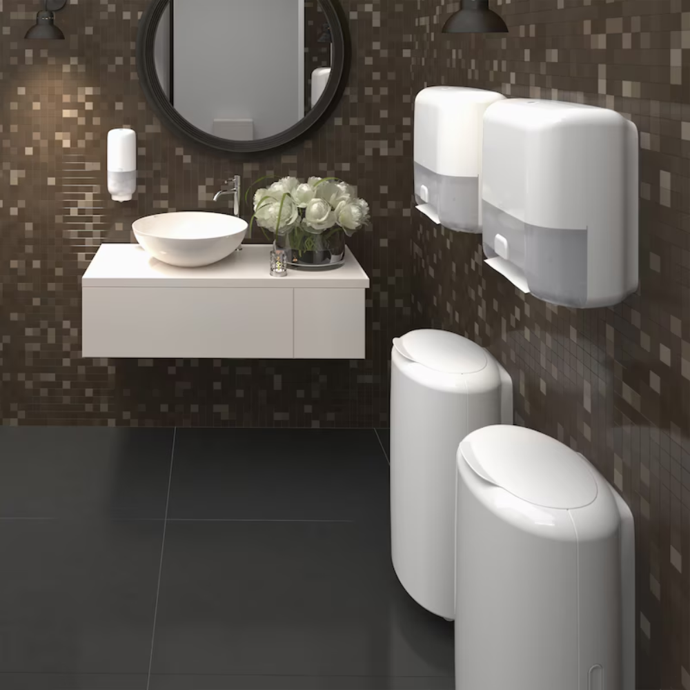 Ein modernes Badezimmer mit dunklen Mosaikfliesen zeigt einen runden Spiegel über einem weißen Aufsatzwaschbecken. Rechts davon sorgen zwei Tork Matic® 551100 Sensorspender für Rollenhandtücher Elevation H1 von TORK – Essity Professional Hygiene Germany GmbH für berührungsloses Händetrocknen und Einzelblattausgabe. Darunter stehen zwei weiße Abfalleimer und ein Strauß weißer Blumen verleiht der Waschtischplatte eine dekorative Note.