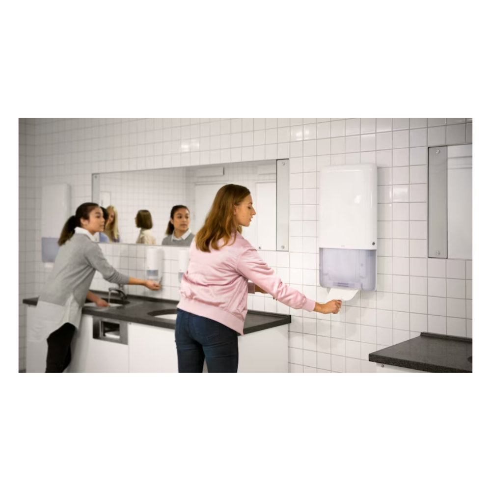 Zwei Frauen stehen in einem Badezimmer mit weiß gefliesten Wänden und großen Spiegeln. Eine benutzt ein Waschbecken auf der linken Seite, während die andere mühelos ein Endlos-Handtuch aus einem Tork PeakServe® 552500 Spender für Endlos-Handtücher Elevation H5 von TORK – Essity Professional Hygiene Germany GmbH zieht, der sich auf der rechten Seite befindet. Die graue Waschtischplatte ergänzt die helle Beleuchtung in diesem stark frequentierten Waschraum.