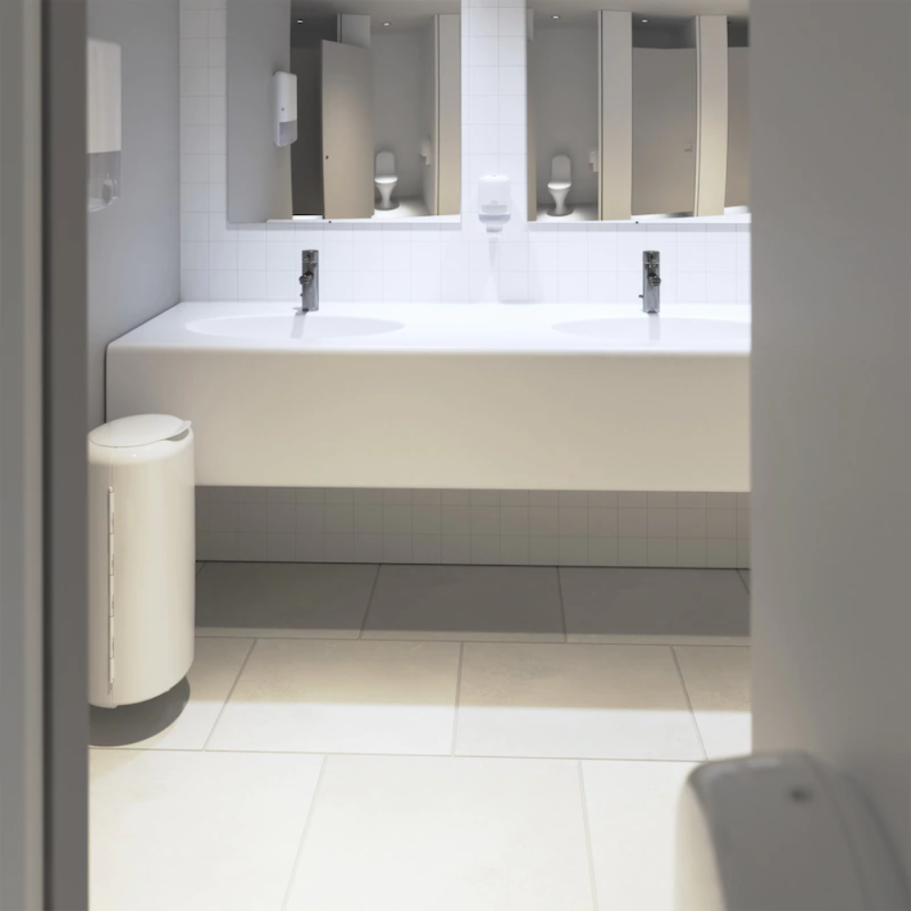 Das moderne öffentliche Badezimmer verfügt über eine elegante weiße Waschtischplatte mit zwei Waschbecken und Chromarmaturen. Links steht ein weißer Mülleimer, während über dem gefliesten Boden Spiegel hängen. An der Wand sind Tork Xpress® 552000 Seifenspender von Essity Professional Hygiene Germany GmbH angebracht, und Multifold Handtücher erhöhen den Komfort der Besucher.