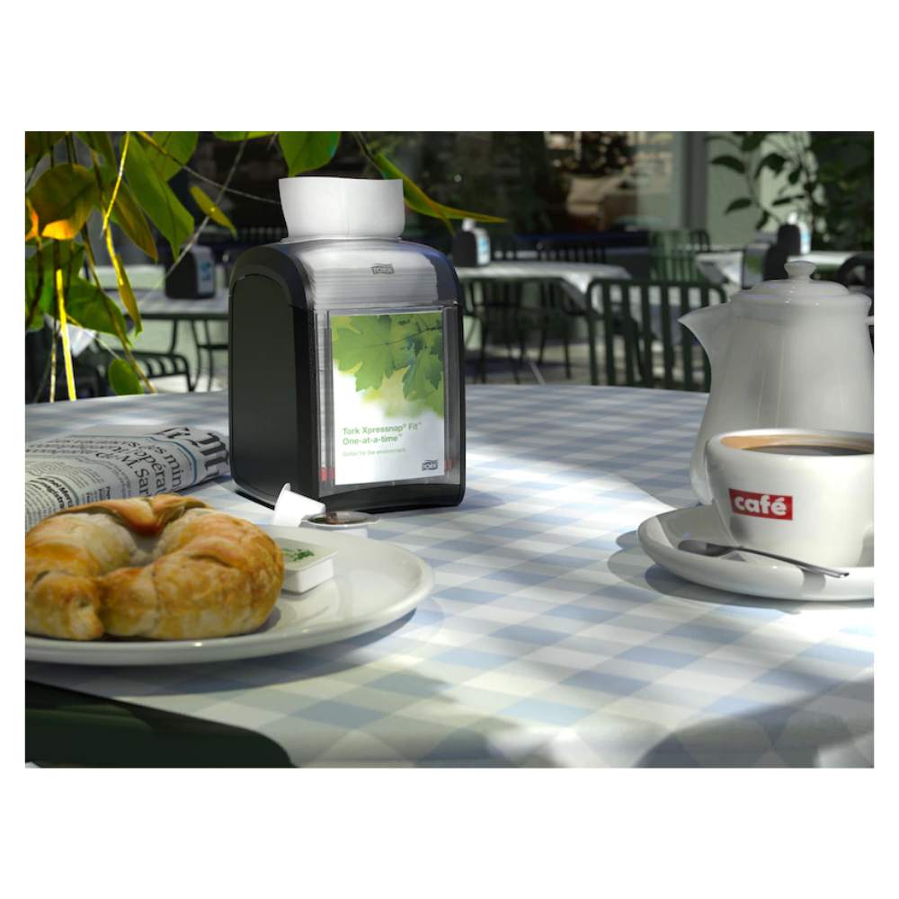 Ein Tisch in einem Café, geschmückt mit einer blau-weiß karierten Tischdecke, ist elegant für eine Person gedeckt. Auf dem Tisch stehen ein Croissant, eine Tasse Kaffee in einer „Café“-Tasse, eine Teekanne und eine Zeitung. Der Tork Xpressnap Fit® 272900 Tischspender für Servietten von TORK – Essity Professional Hygiene Germany GmbH steht hübsch daneben. Sonnenlicht fällt durch die Pflanzen in der Nähe.
