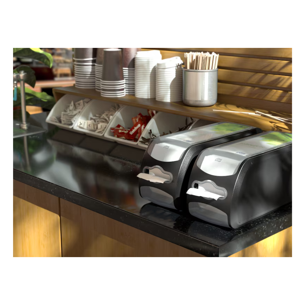 Eine Kaffeestation ist mit einem Tork Xpressnap Fit® 272901 Thekenspender für Servietten von TORK – Essity Professional Hygiene Germany GmbH ausgestattet, der über zwei Serviettenspender verfügt. Außerdem sind ordentlich gestapelte Pappbecher, Deckel, Rührstäbchen und Gewürze wie Zucker- und Sahnebeutel in Fächern auf einer eleganten schwarzen Theke angeordnet.