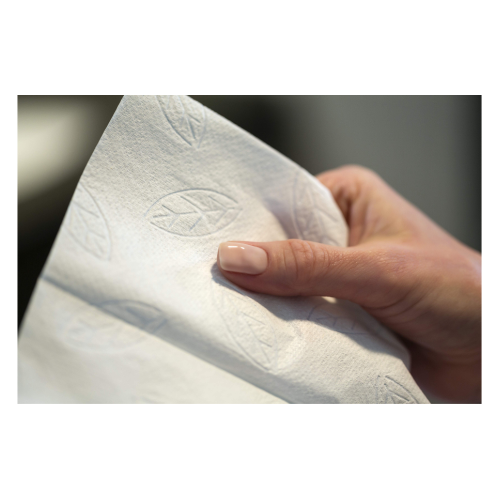 Eine Hand hält ein hochwertiges weißes Tork 100278 extraweiche Zickzack-Handtücher Premium H3 2-lagig Papierhandtuch, geprägt mit Blattmuster. Daumen und Zeigefinger umfassen dieses Hygieneutensil von TORK – Essity Professional Hygiene Germany GmbH. Der Hintergrund ist unscharf, was die Eleganz des Handtuchs hervorhebt.