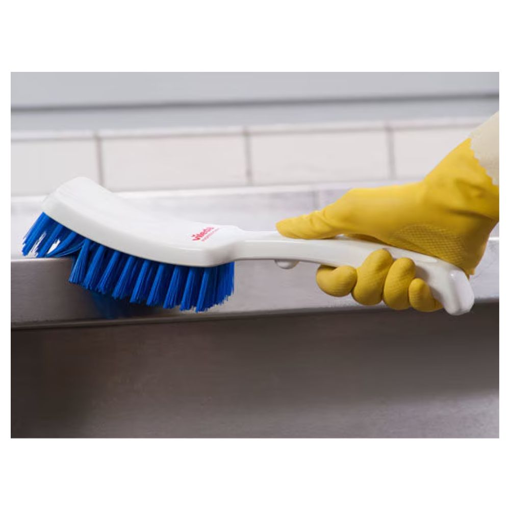 Eine Person mit gelben Gummihandschuhen schrubbt mit einer Vileda Professional Stielbürste mit kurzem Griff blau – 30 cm der Vileda GmbH eine graue Oberfläche, wobei eine geflieste Wand einen neutralen Hintergrund bietet.