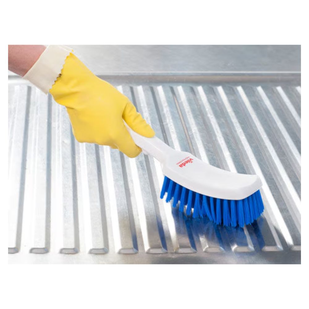 Eine Person mit einem gelben Gummihandschuh verwendet eine Vileda Professional Stielbürste mit kurzem Griff blau – 30 c| Packung (1 Stück) der Vileda GmbH mit hochwertigen Borsten zum Reinigen einer geriffelten Metalloberfläche.