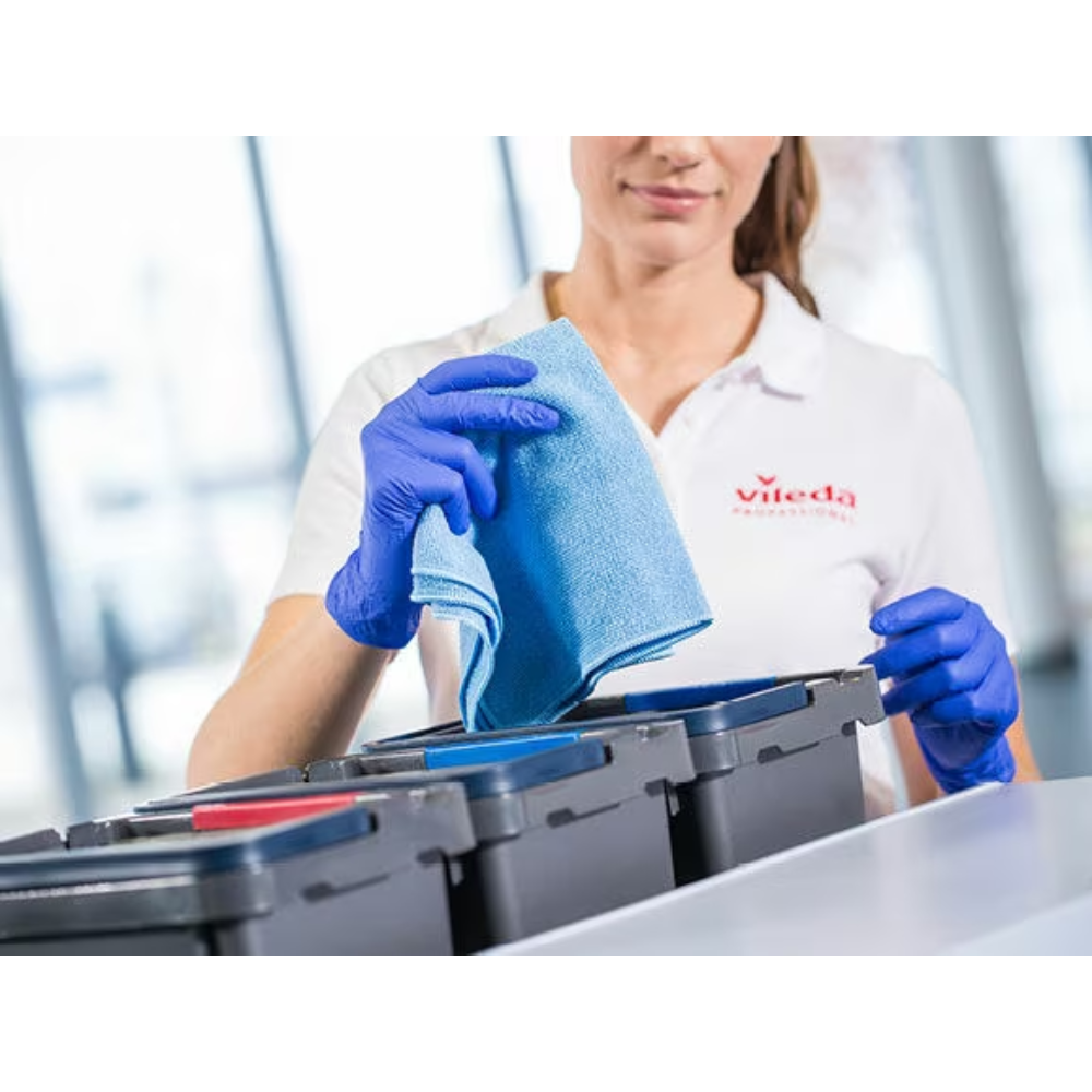 Eine Person mit weißem Hemd und blauen Handschuhen hält ein blaues Vileda Professional r-MicroTuff Swift Microfasertuch der Vileda GmbH über eine Reihe von Behältern mit verschiedenfarbigen Deckeln; die Szene verdeutlicht die effiziente Reinigungsaufgabe mithilfe des Vier-Farb-Systems zur Ordnung.