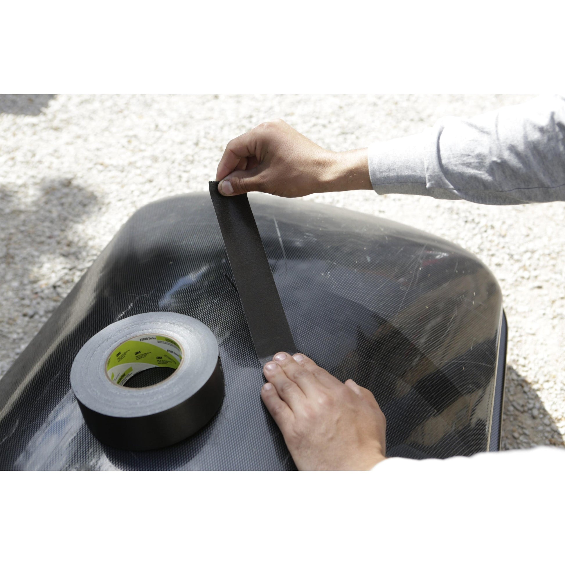 Eine Person im langärmeligen Hemd klebt das Scotch® Extremium™ Ultra Hochleistungs-Klebeband DT17 (10 m x 24 mm) der 3M Deutschland GmbH auf eine glatte, dunkle Oberfläche im Freien, um den Einsatz für extreme Anforderungen zu zeigen, wobei die Klebebandrolle in der Nähe ist.