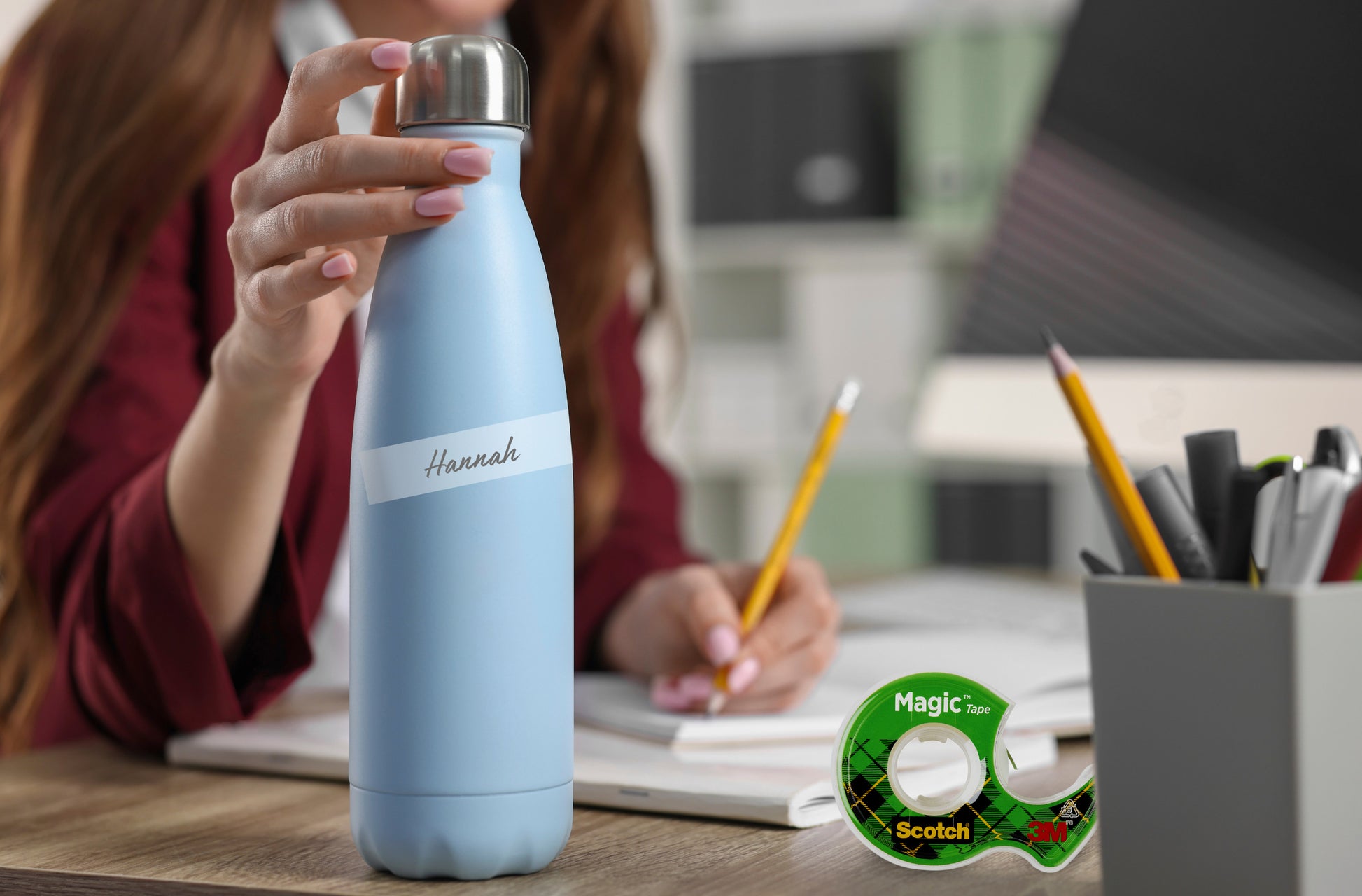 Eine Person an einem Schreibtisch hält eine hellblaue Wasserflasche mit der Aufschrift "Hannah"; daneben liegen Büromaterialien, ein Bleistift und eine Scotch® Magic™ Unsichtbares Klebeband Nachfüllpackung (1 Rolle, 19 mm x 25 m) der 3M Deutschland GmbH.