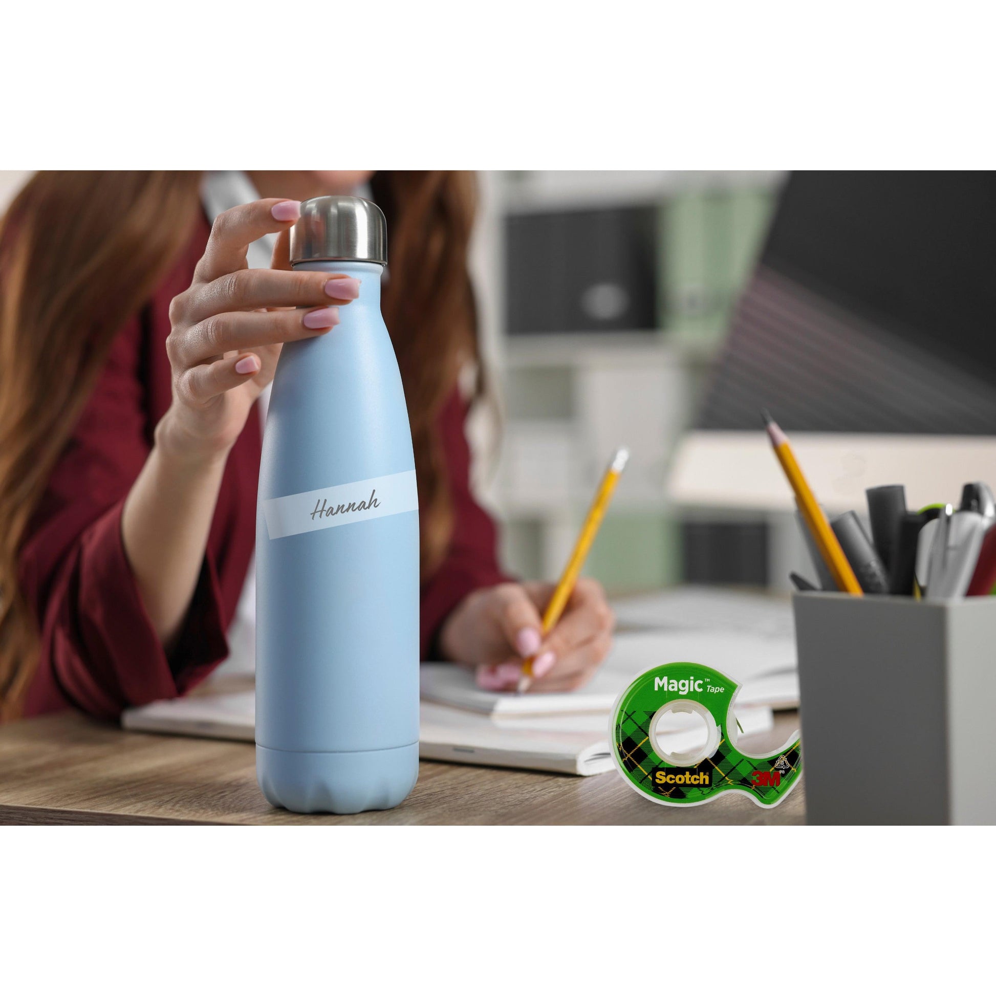 Eine Person hält eine hellblaue Wasserflasche mit der Aufschrift "Hannah" an einem Schreibtisch mit Bleistiften, Büromaterial und einer Packung Scotch® Magic™ Unsichtbares Klebeband (2 Rollen, 19 mm x 7,5 m + 1 Handabroller gratis) von 3M Deutschland GmbH.