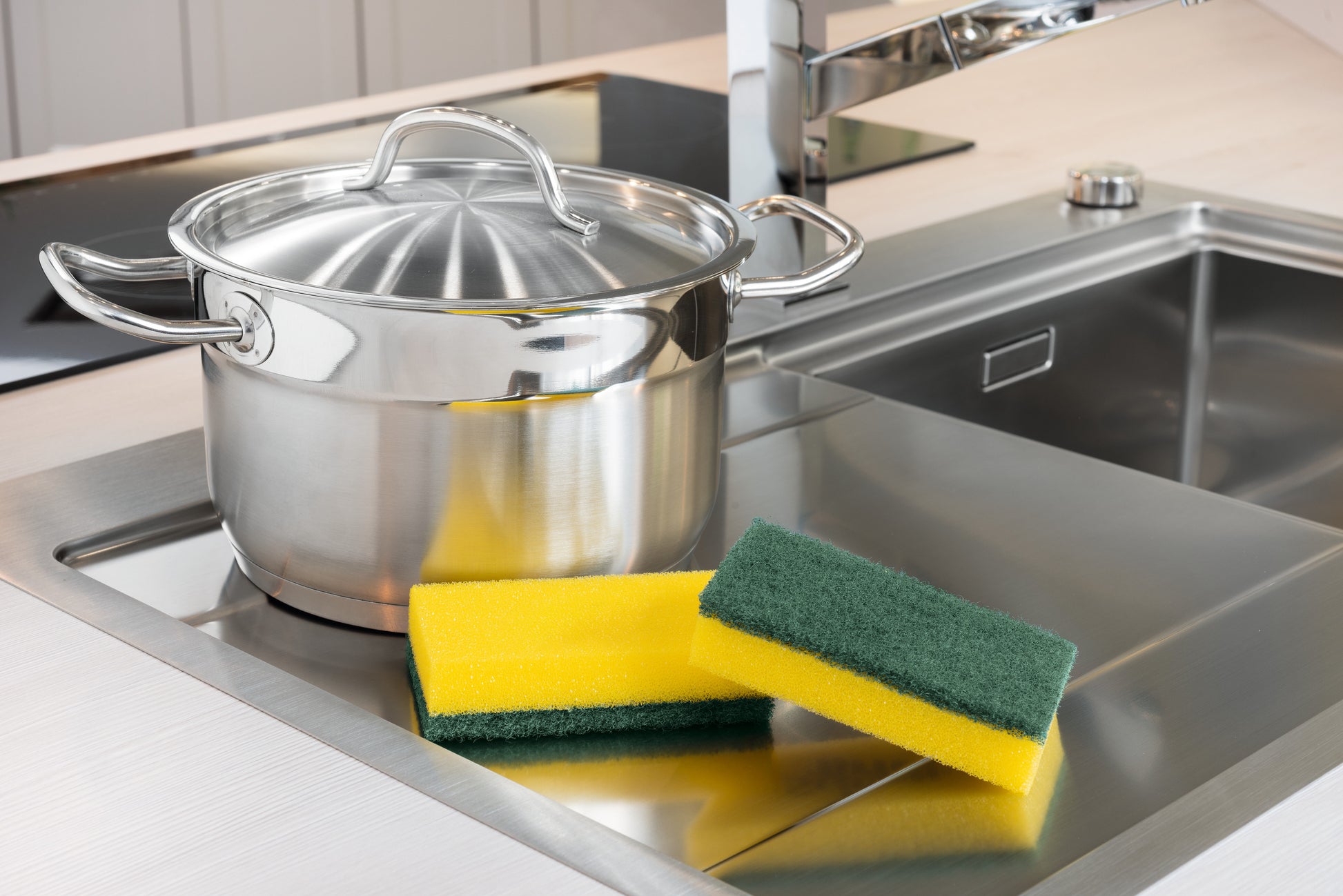 Ein Edelstahltopf mit Deckel steht auf einer Metallspüle neben zwei gelb-grünen Scotch-Brite® Classic Reinigungsschwamm XXL der 3M Deutschland GmbH; im Hintergrund sind auch der Wasserhahn und die Arbeitsplatte zu sehen.