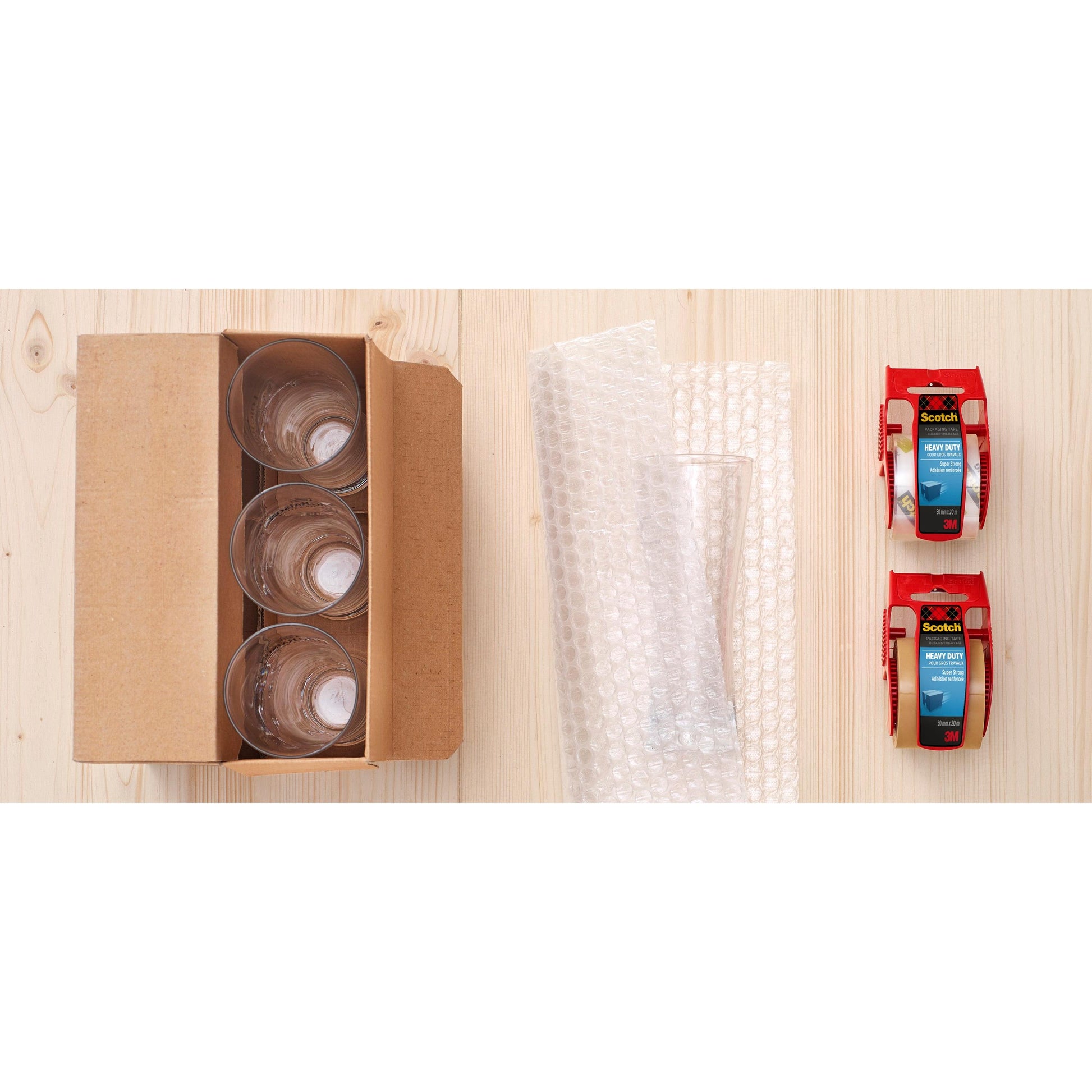 Ein Karton mit vier Glasbechern, ein Blatt Luftpolsterfolie, ein leeres Glas und eine Rolle Scotch® Extra starkes Verpackungsklebeband (Braun, 48 mm x 20,3 m) der 3M Deutschland GmbH stehen fein säuberlich auf einer hellen Holzfläche.