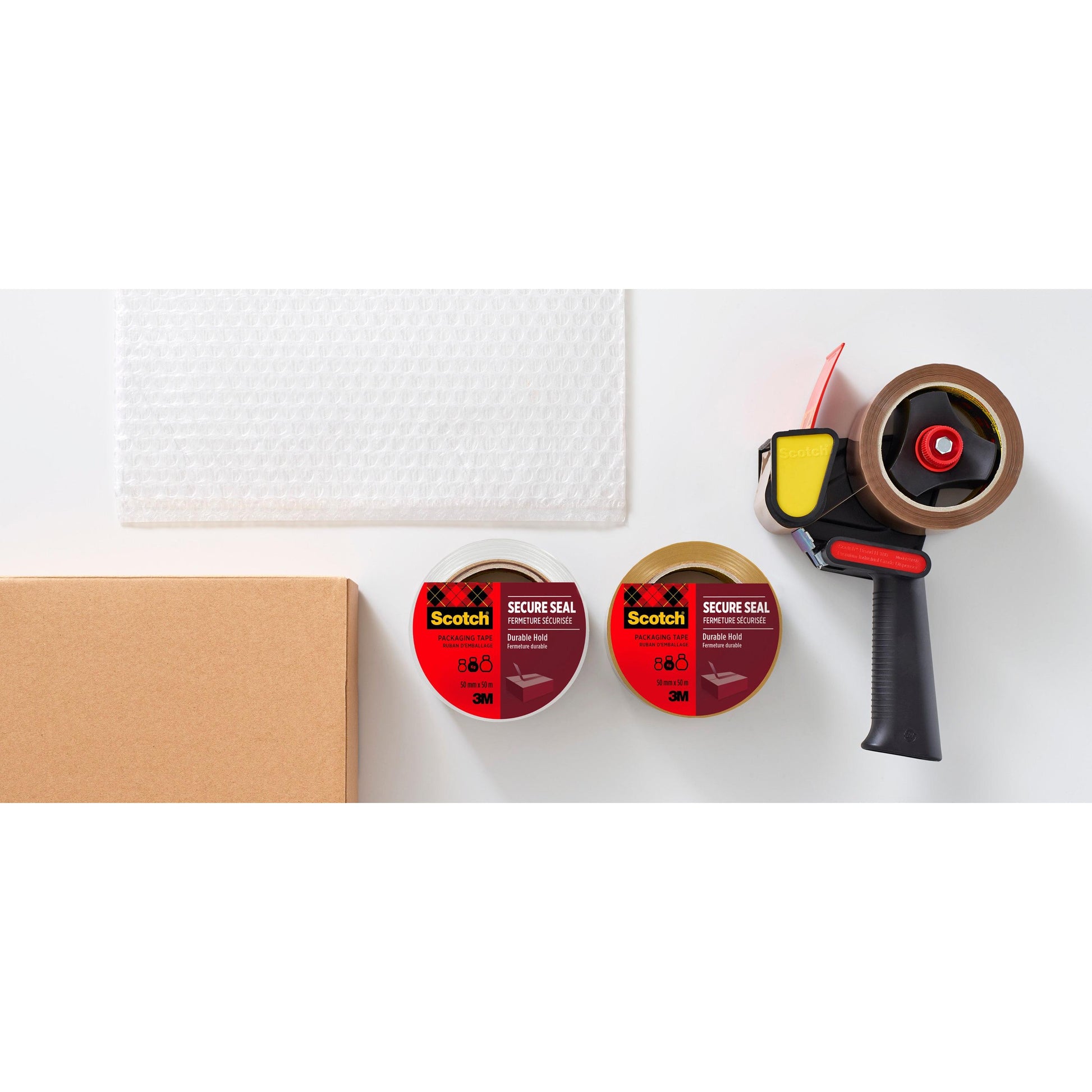 Eine Rolle Luftpolsterfolie, ein Karton, zwei Rollen 3M Deutschland GmbH Scotch® Verpackungsklebeband (50 mm x 66 m) und ein schwarzer Klebebandabroller sind auf einer weißen Fläche angeordnet.