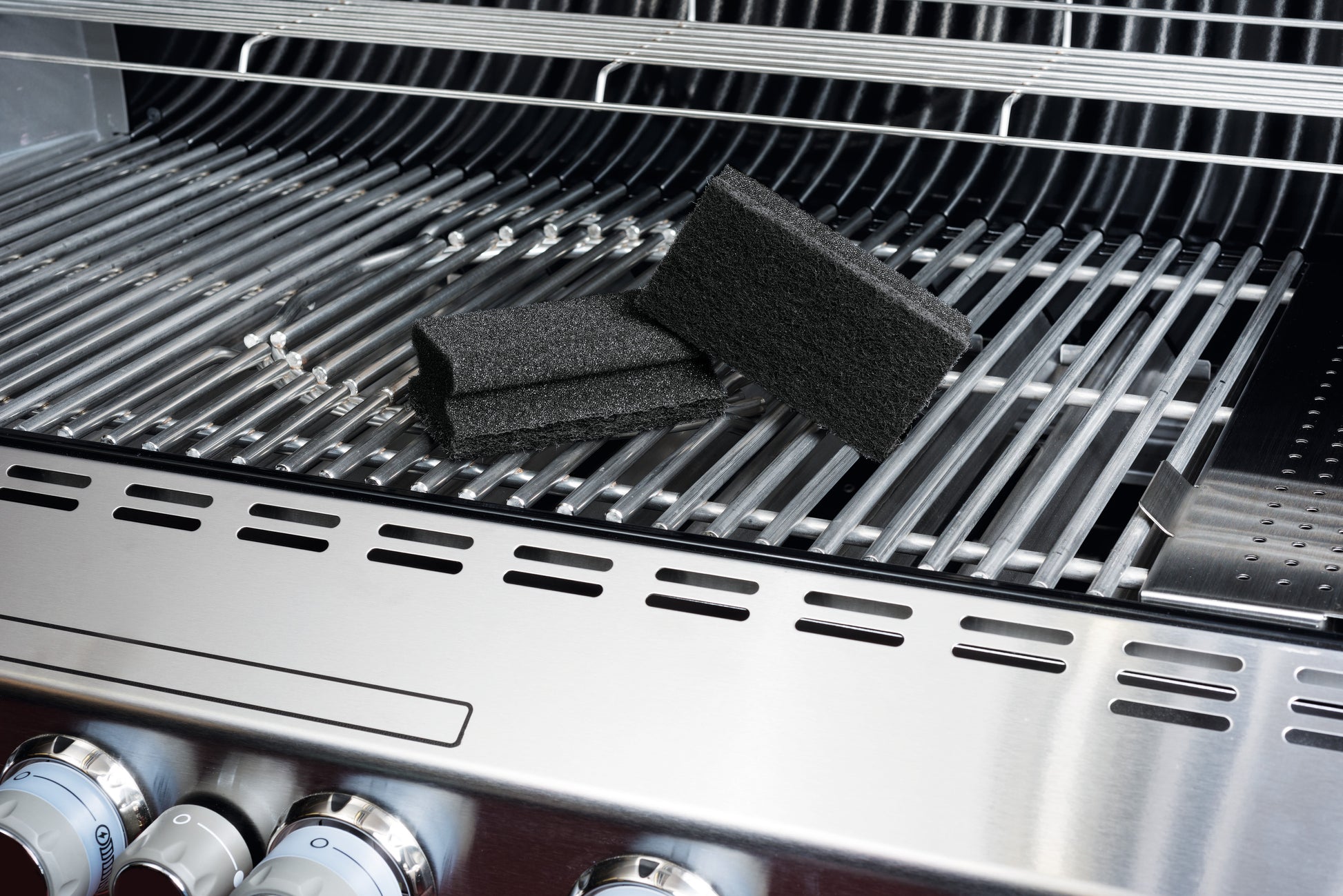 Auf einem sauberen Edelstahl-Gasgrill liegen zwei Scotch-Brite® Grill-Griffschwämme von 3M Deutschland GmbH auf den Grillrosten, wobei die Bedienknöpfe an der Vorderseite sichtbar sind. Jeder Schwamm wird in einer Packung mit einem Stück geliefert.