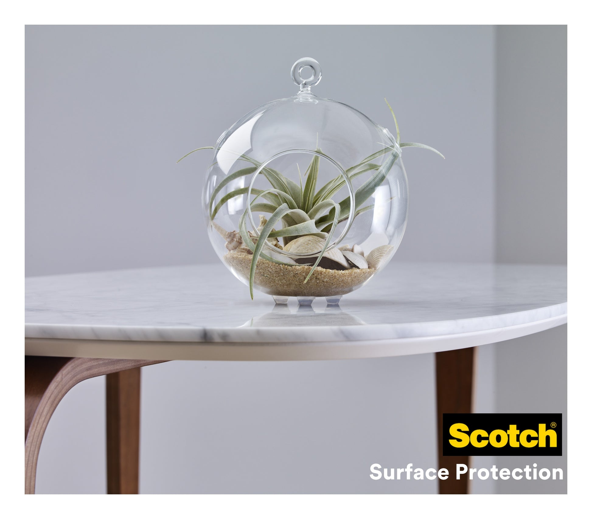Ein Glasterrarium mit Luftpflanzen und Sand steht auf einer weißen Marmortischplatte mit Holzbeinen. In der Ecke des Tisches steht das Logo der 3M Deutschland GmbH und "Scotch® Stoßabsorbierende Elastikpuffer, selbsthaftend, transparent".
