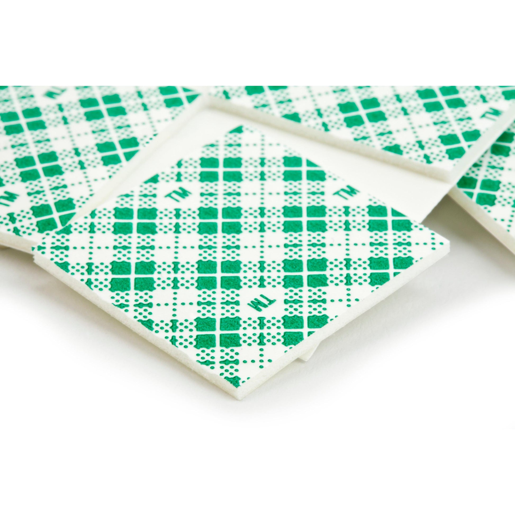 Nahaufnahme von 3M Deutschland GmbH Scotch-FIX™ doppelseitigen Montagequadraten (25 mm x 25 mm, 16 Stück/Packung) in Einzelverpackungen mit grünem Karomuster und verstreuten Buchstaben auf der Verpackung.