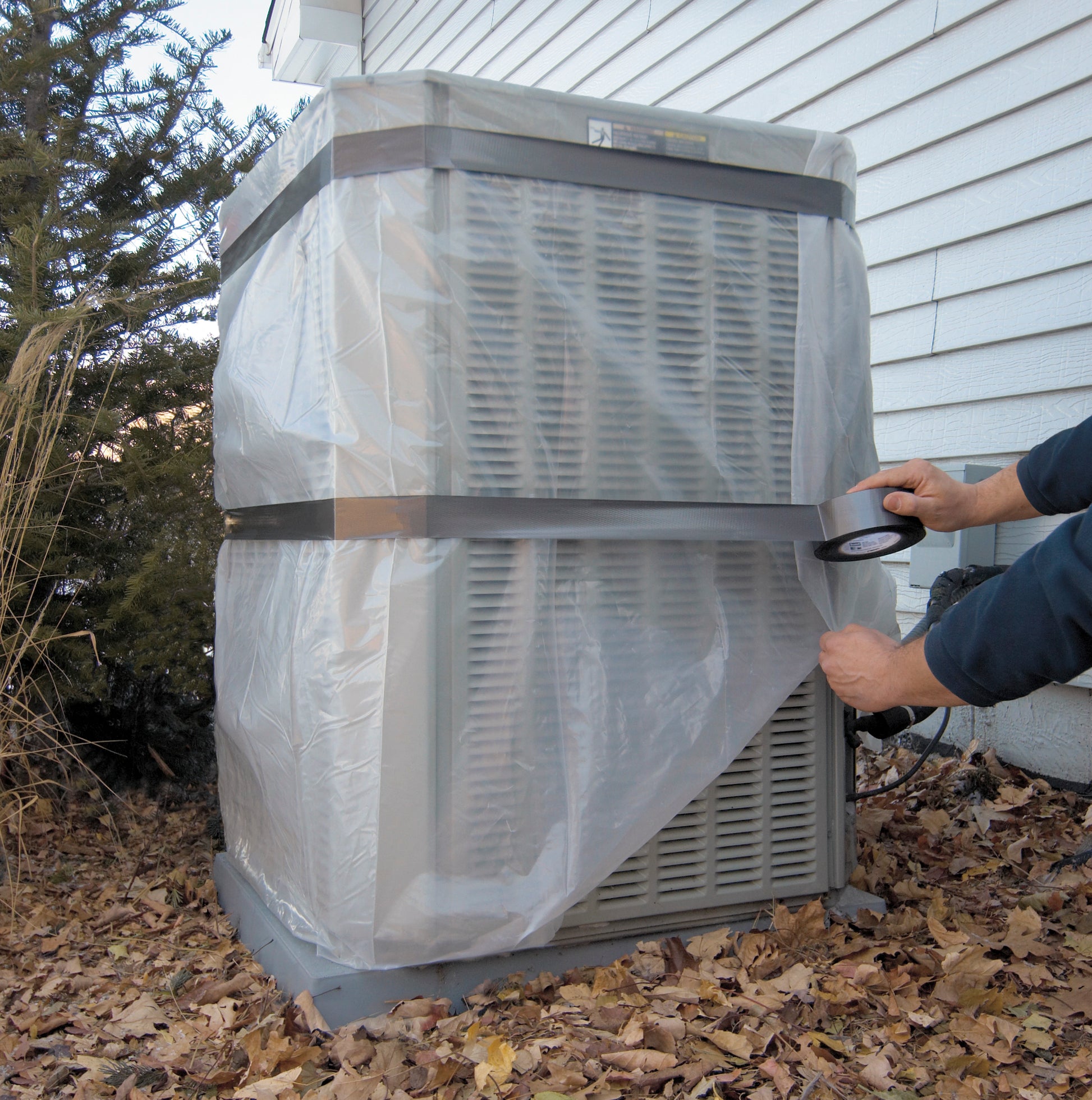 Jemand sichert ein großes Außenklimagerät neben einem Haus mit dem 3M Scotch® Extremium™ All Weather Hochleistungs-Klebeband (27 m x 48 mm) und Plastikfolie inmitten von trockenen Blättern und Pflanzen.