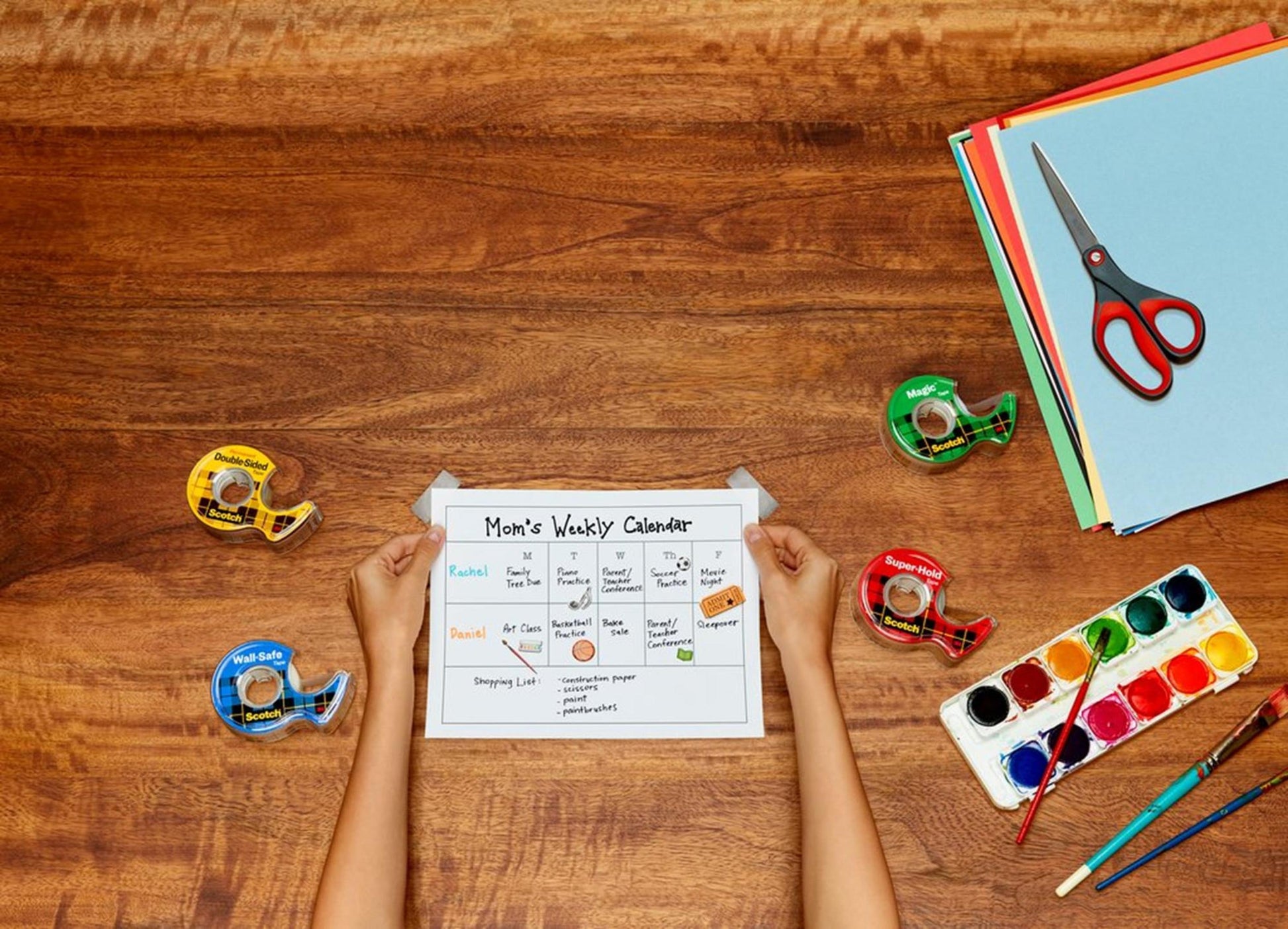Eine Person hält einen "Mom's Weekly Calendar" auf einem Holztisch mit vier Klebebandabrollern, farbigem Papier, einem Aquarellset mit Pinsel und einer Scotch™ Präzisionsschere rot von 3M Deutschland GmbH.