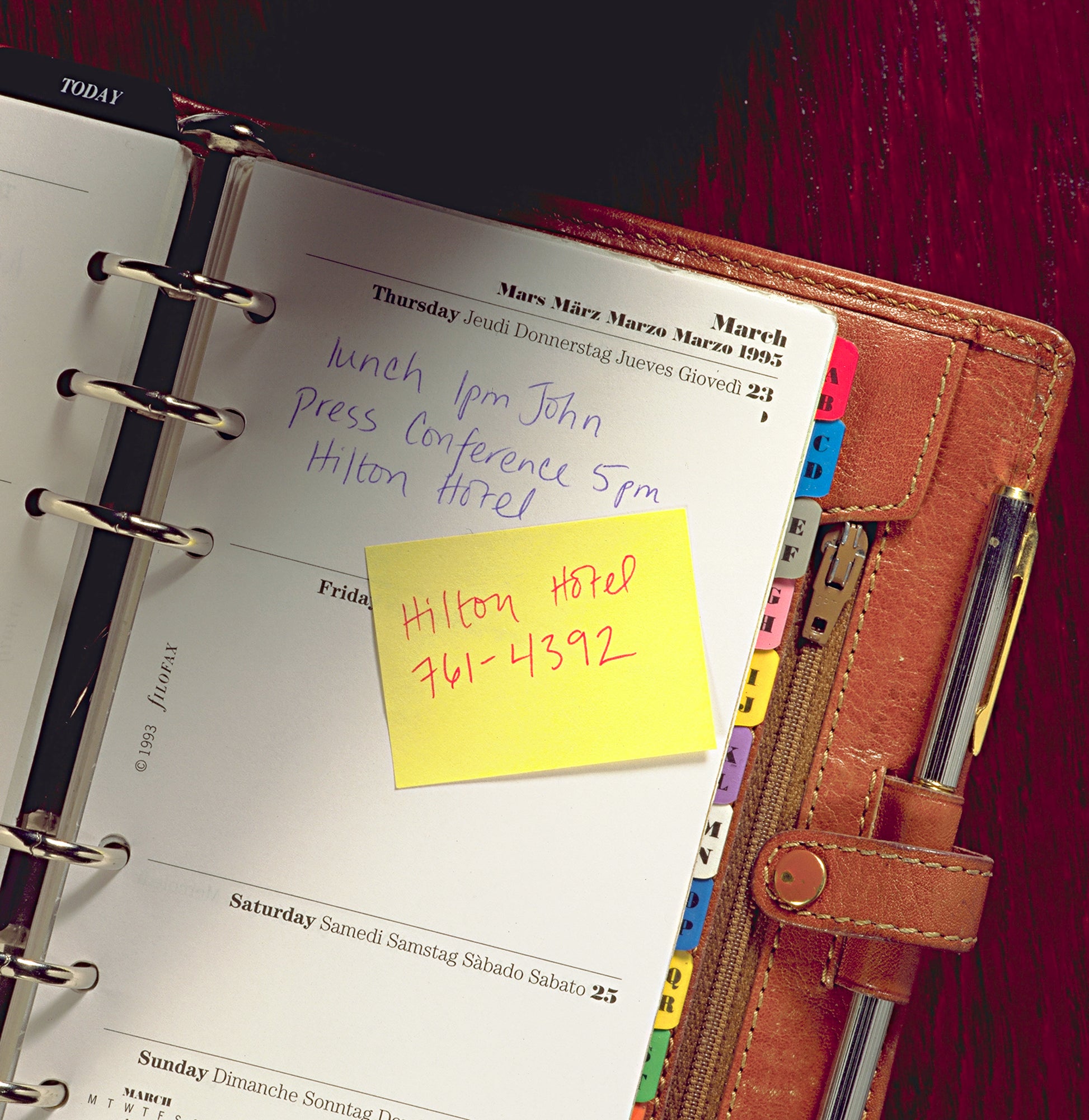 Ein aufgeschlagener ledergebundener Planer zeigt handschriftliche Donnerstagsnotizen: "Mittagessen 13 Uhr John", "Pressekonferenz 17 Uhr Hilton Hotel". Auf einem gelben Post-it® Recycling Notes Block der 3M Deutschland GmbH steht: "Hilton Hotel 761-4392". Die Trennblätter sind sichtbar.