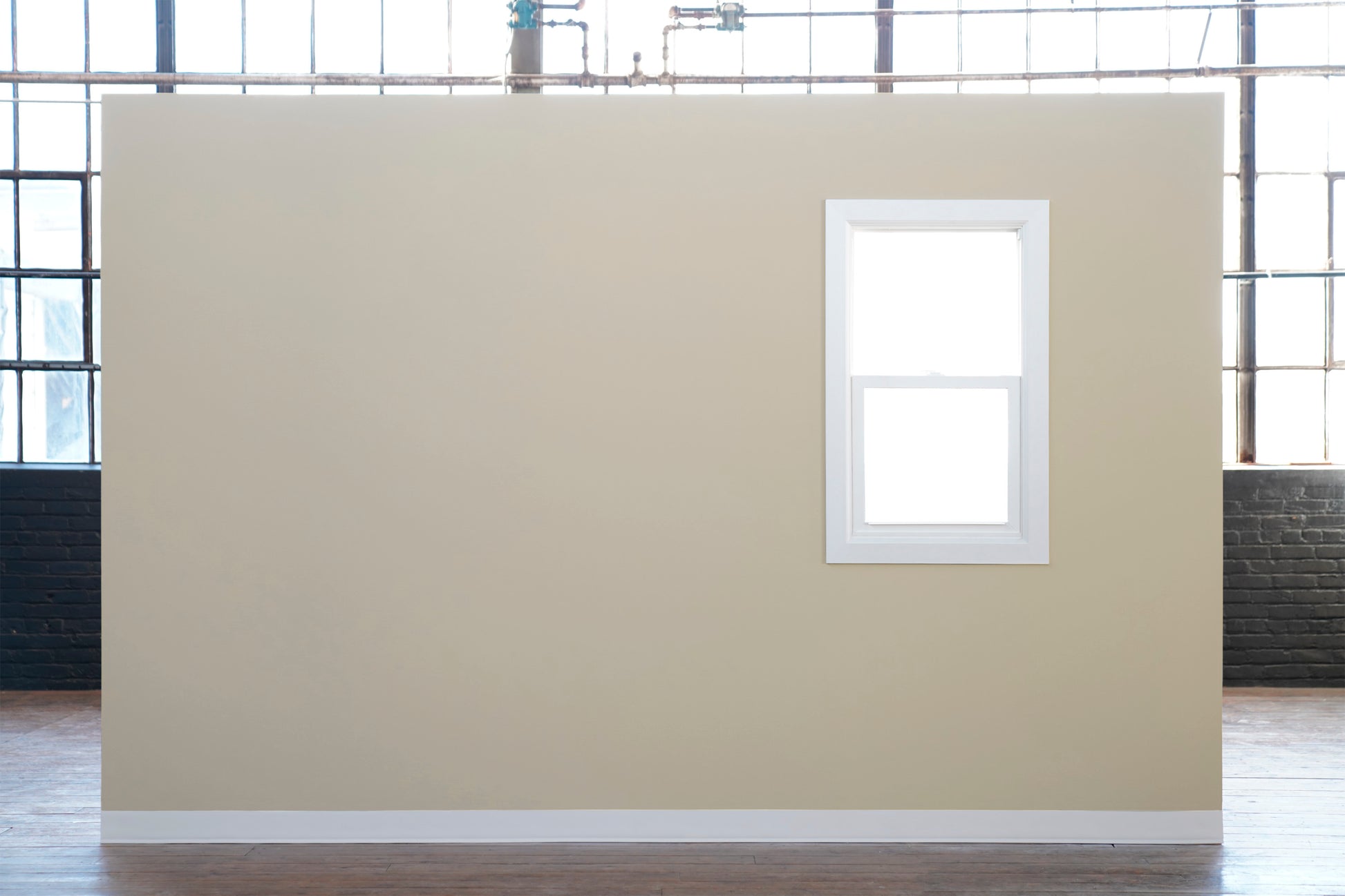 Eine beige Innenwand mit einem weiß gerahmten Fenster steht auf einem Holzboden vor Industriefenstern. Zum präzisen Streichen verwenden Sie Scotch® Classic Abdeckband, 36 mm x 50 m von 3M Deutschland GmbH-70% PEFC zertifiziert (1 Rolle pro Packung).