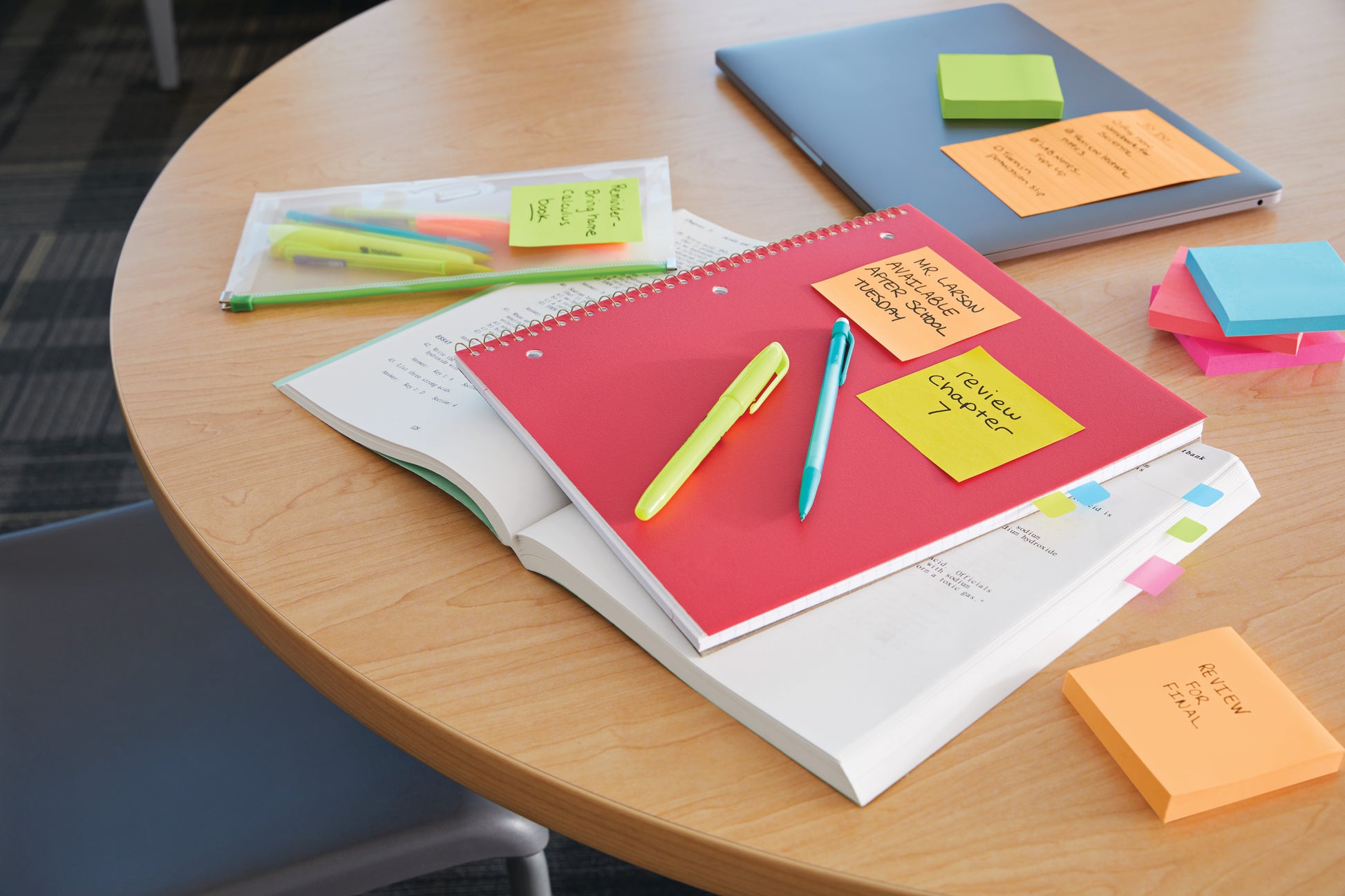 Ein runder Holztisch mit einem geschlossenen Laptop, Notizbüchern, Stiften in einer Plastiktasche, Textmarkern, Erinnerungsnotizen und einem Stapel 3M Deutschland GmbH Post-it® Super Sticky Notes Würfel, Gelb (76 mm x 76 mm). Ein Stuhl ist teilweise sichtbar.