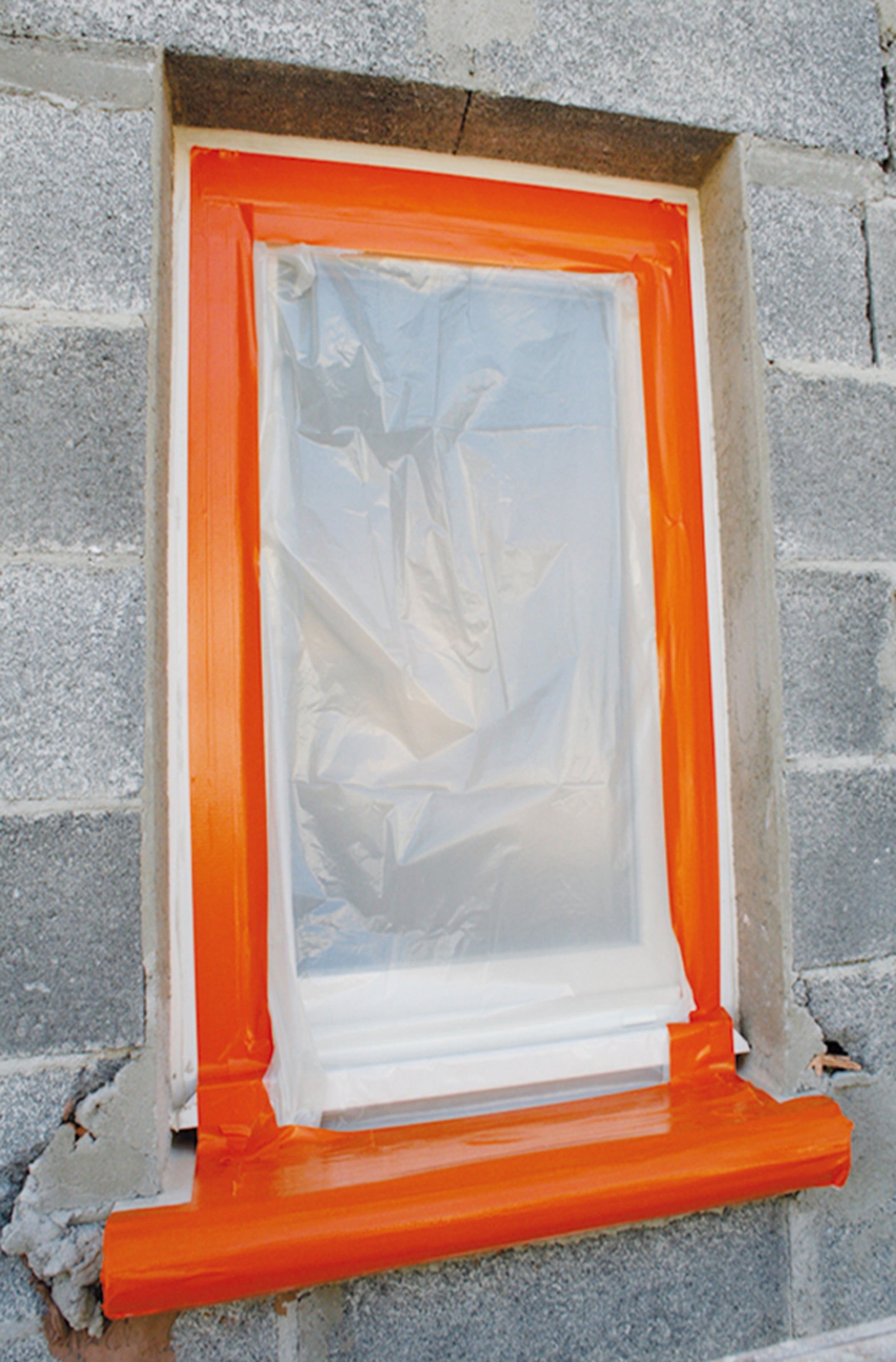 Eine Fensteröffnung in einer Schlackenwand ist mit Kunststoff abgedeckt, gesichert mit 3M Deutschland GmbH Scotch® High Visibility Klebeband, orange (25 m x 48 mm). Die unfertige Wand und der Rahmen weisen an der Basis einige Schäden auf.