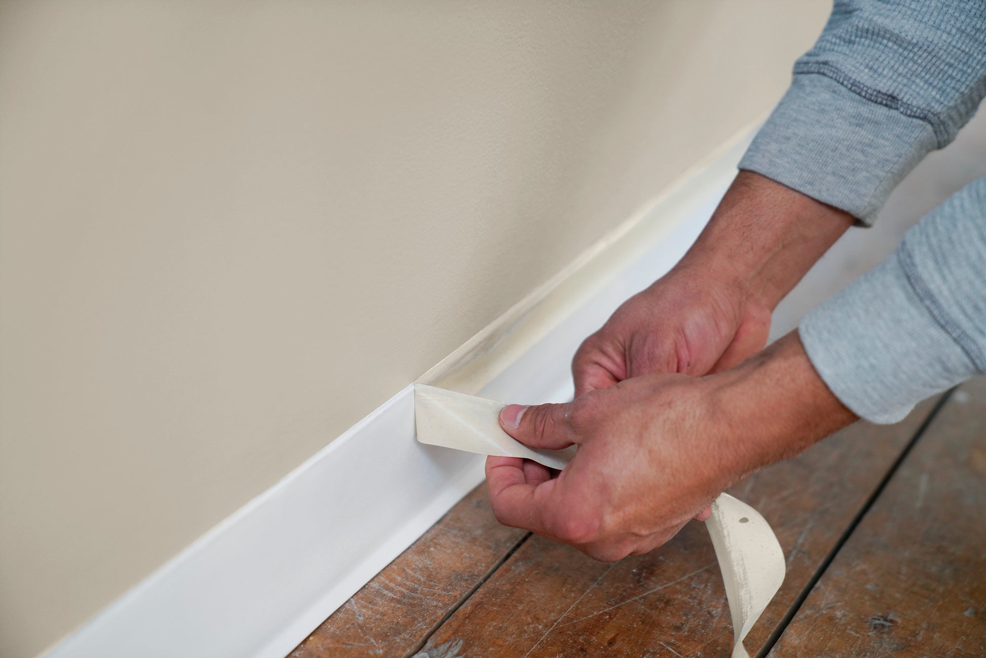 Eine Person in einem grauen langärmeligen Hemd zieht Scotch® Classic Abdeckband (24 mm x 50 m, 3M Deutschland GmbH) von einer weißen Fußleiste entlang einer beigen Wand ab und legt eine saubere Farblinie über dem Holzboden frei.