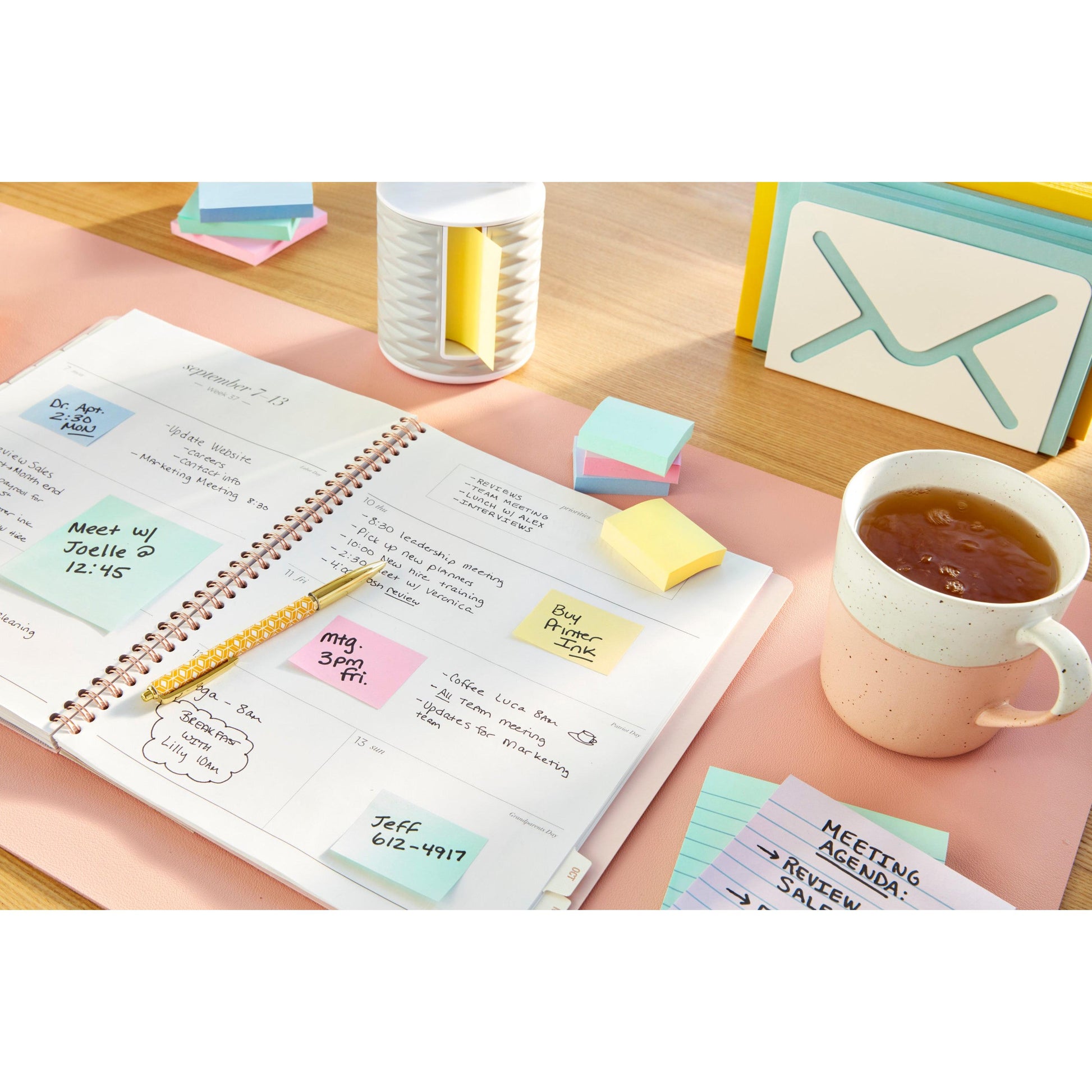 Ein offener Planer mit handschriftlichen Notizen, ein Stift, 3M Deutschland GmbH Post-it® Recycling Notes (76 mm x 127 mm, 100% PEFC), eine Tasse Tee, eine Karteikarte mit der Aufschrift "MEETING AGENDA" und Schreibtischzubehör auf einer hellrosa Schreibtischunterlage.