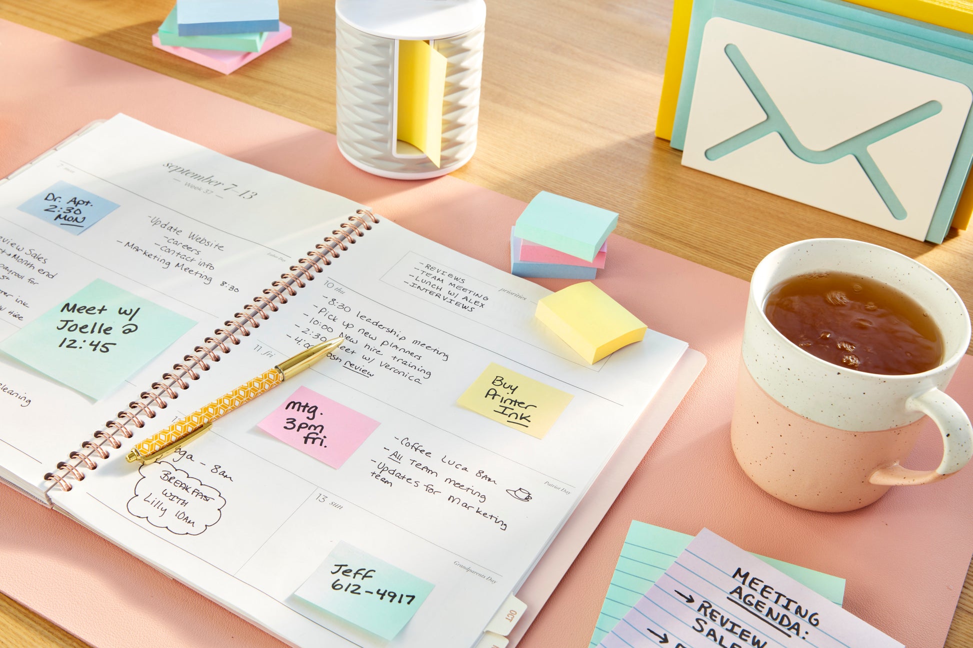 Ein offener Planer zeigt einen Wochenplan auf einem Schreibtisch, umgeben von handschriftlichen Notizen, einem Stift, Büromaterial, kleinen Karten, einer Tasse Tee und Post-it® Recycling Notes (38 mm x 51 mm) der 3M Deutschland GmbH.
