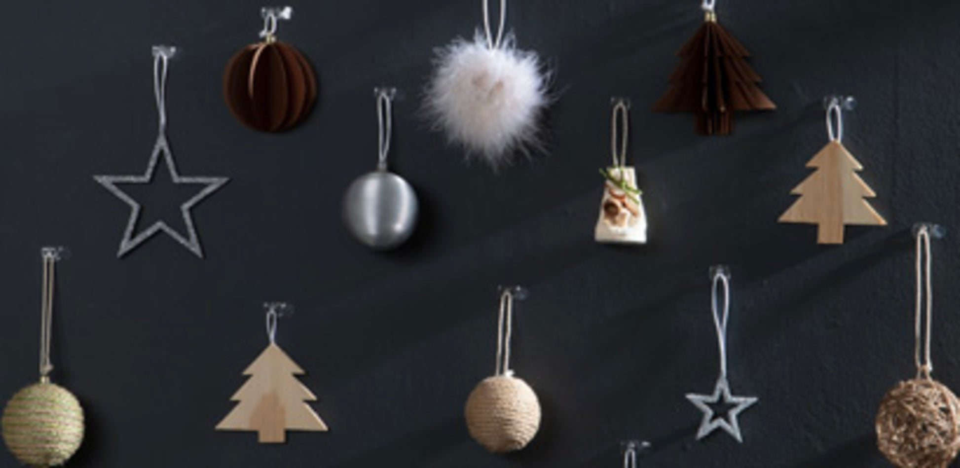 Weihnachtsschmuck - Sterne, Bäume, Kugeln und eine flauschige weiße Dekoration aus Holz, Metall, Schnur und Stoff - hängen an einer dunklen Wand mit Command™ Dekorations-Clips (transparent) von 3M Deutschland GmbH.