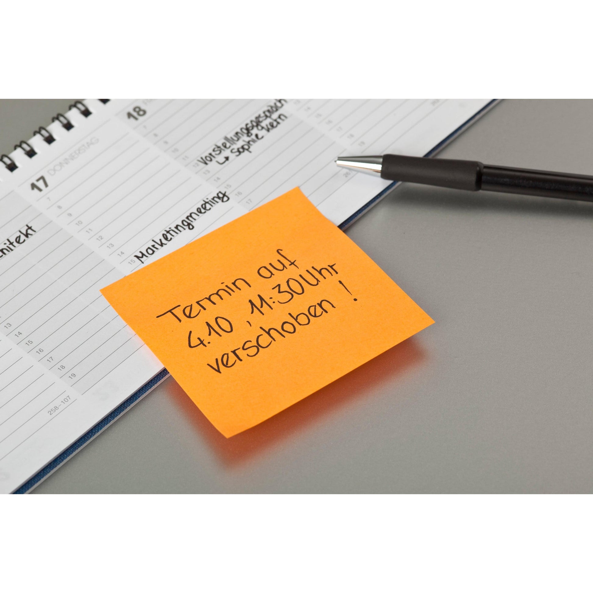 Ein schwarzer Stift, ein aufgeschlagener Planer und ein Post-it® Zettel der 3M Deutschland GmbH (76 mm x 76 mm, 100 Blatt/Block, 100% PEFC) mit der Aufschrift "Termin am 4.10., 11:30 Uhr verschoben!" auf den Planer gelegt.
