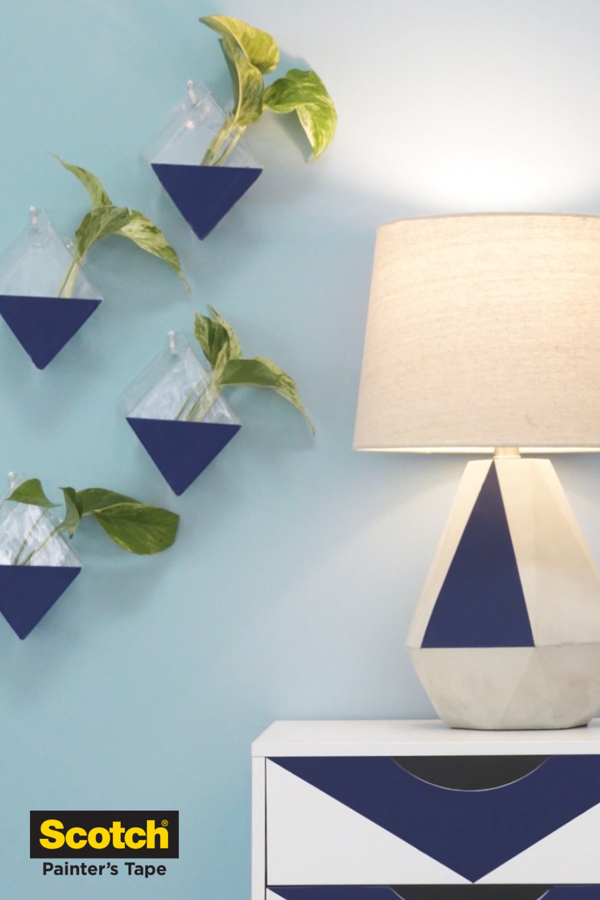 Eine weiße und blaue geometrische Tischlampe steht auf einer passenden Kommode neben einer hellblauen Wand mit drei dreieckigen Pflanzgefäßen mit Grünpflanzen. Das Scotch® Malerabdeckband für empfindliche Oberflächen by 3M Deutschland GmbH Logo befindet sich in der unteren linken Ecke.