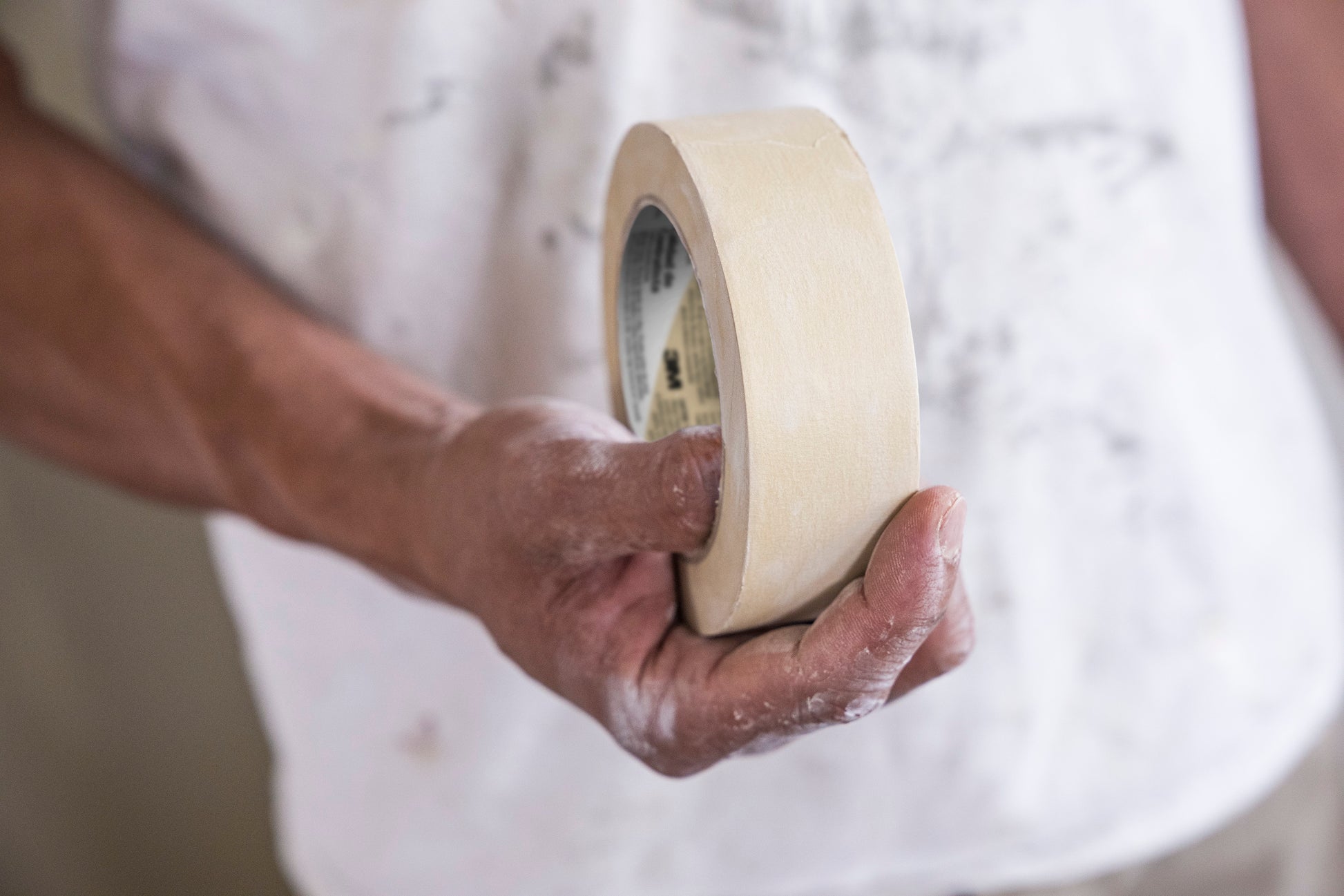 Eine Person mit farbverschmierten Händen und einem verschmierten weißen Hemd hält eine Rolle Scotch® Classic Abdeckband (36 mm x 50 m) der 3M Deutschland GmbH - ideal für Maler- und Renovierungsarbeiten.