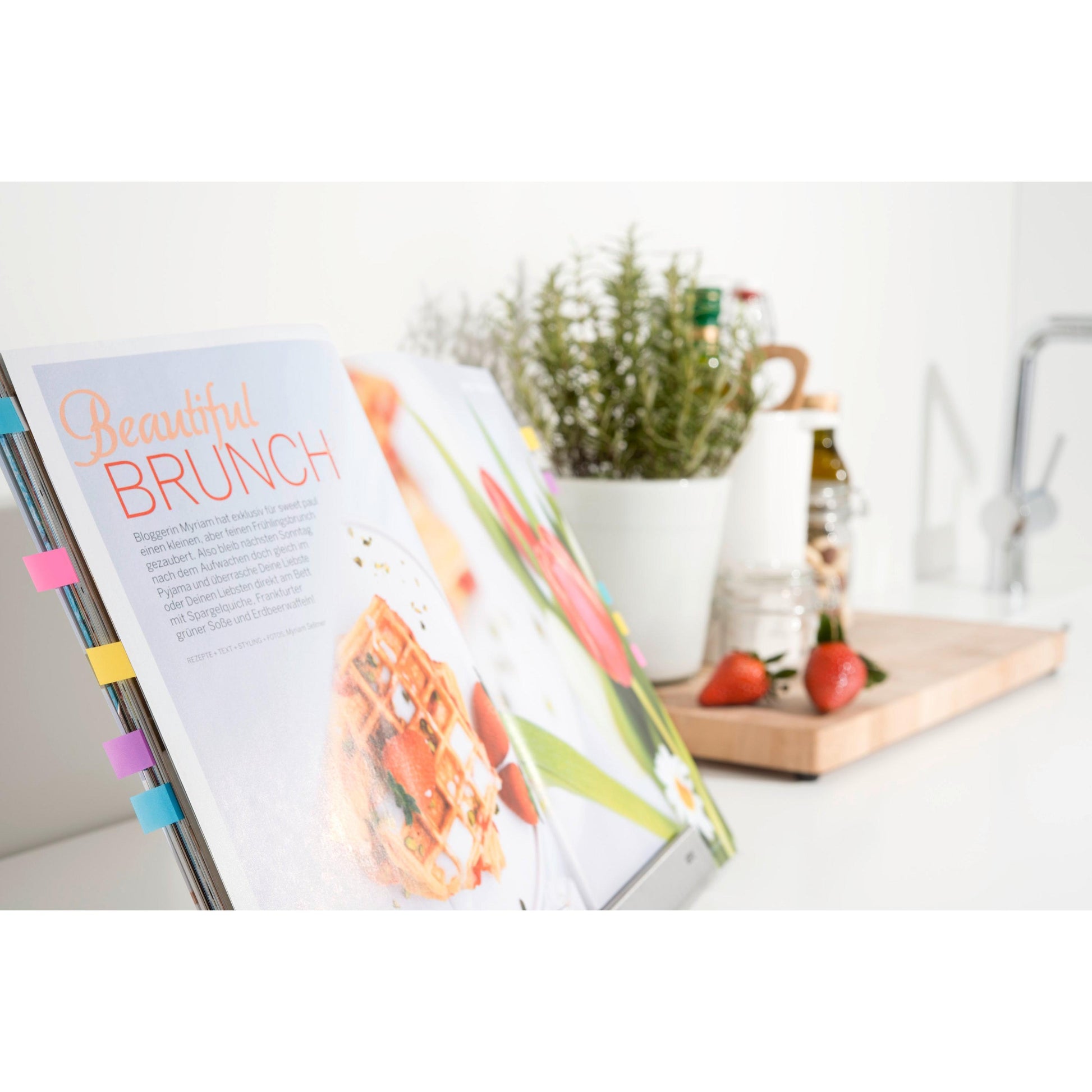 Ein mit Post-it® Index Haftstreifen Mini (11,9 x 43,2 mm, 100/Spender) beschriftetes Kochbuch der 3M Deutschland GmbH zeigt ein Brunch-Rezept; dahinter stehen ein weißer Kräutertopf, Erdbeeren auf einem Brett und Küchenutensilien auf einer sonnenbeschienenen Theke.