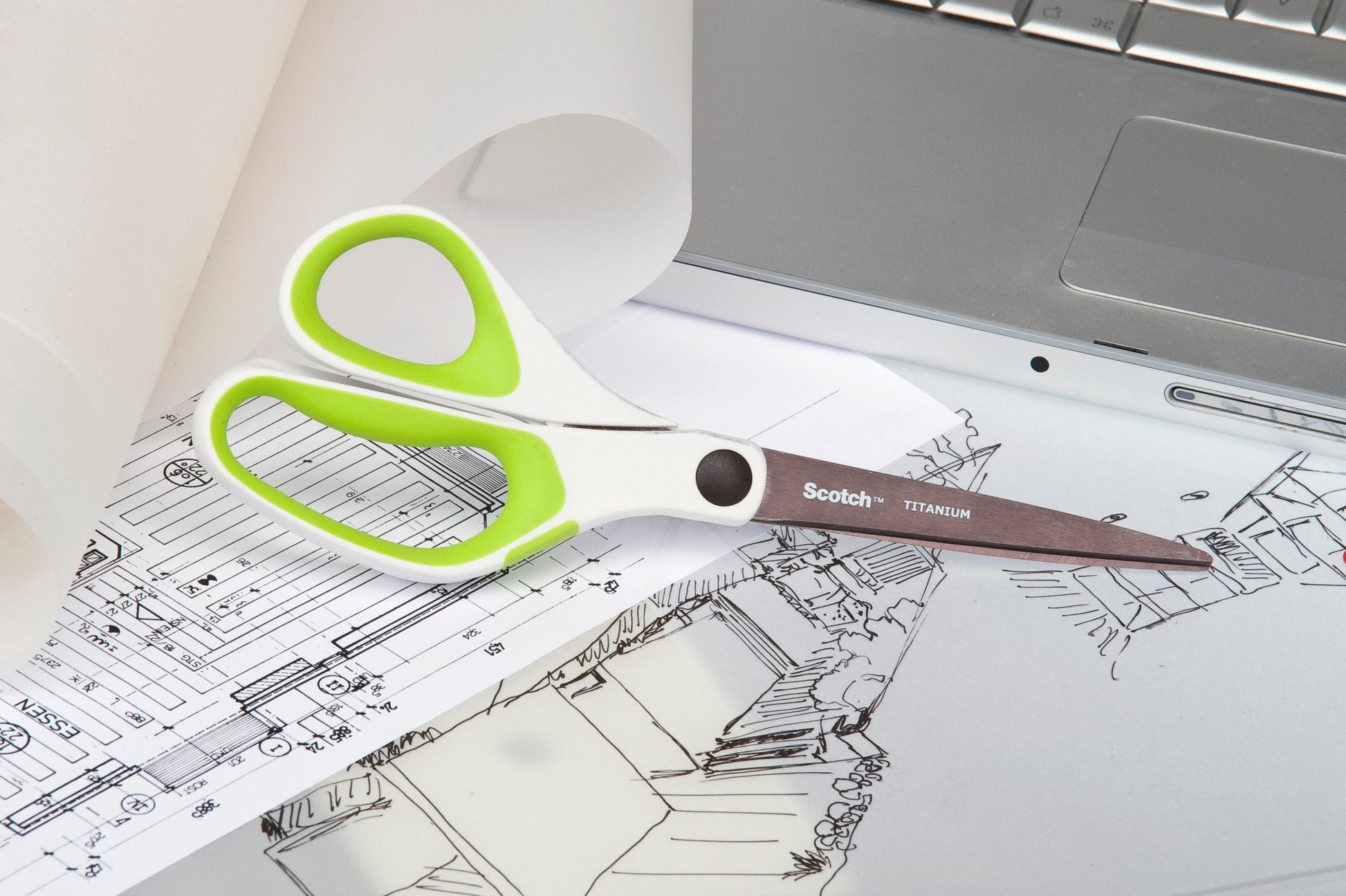 Ein Paar Scotch™ Titanschere, 20 cm von 3M Deutschland GmbH, mit weißen und grünen Griffen, ruht auf Architekturzeichnungen neben einem silbernen Laptop und einer Rolle Transparentpapier.