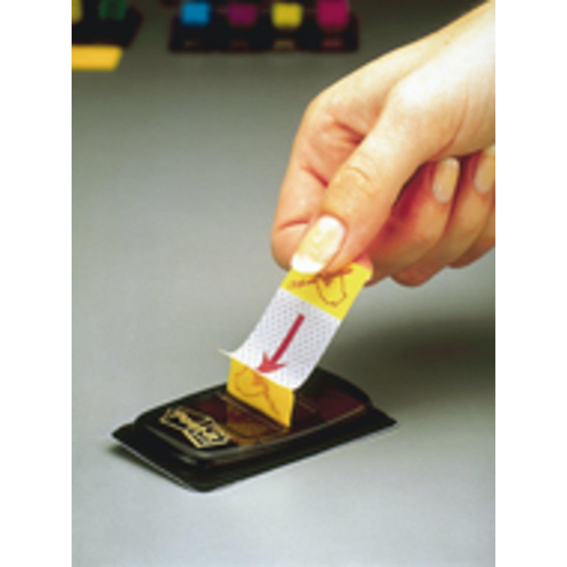 Eine Hand hebt einen gelben Post-it® Index Haftstreifen (25,4mm x 43,2mm, 50 Stück) von 3M Deutschland GmbH aus seinem schwarzen Spender auf einer grauen Fläche. Die farbigen Würfel im Hintergrund sind leicht unscharf.