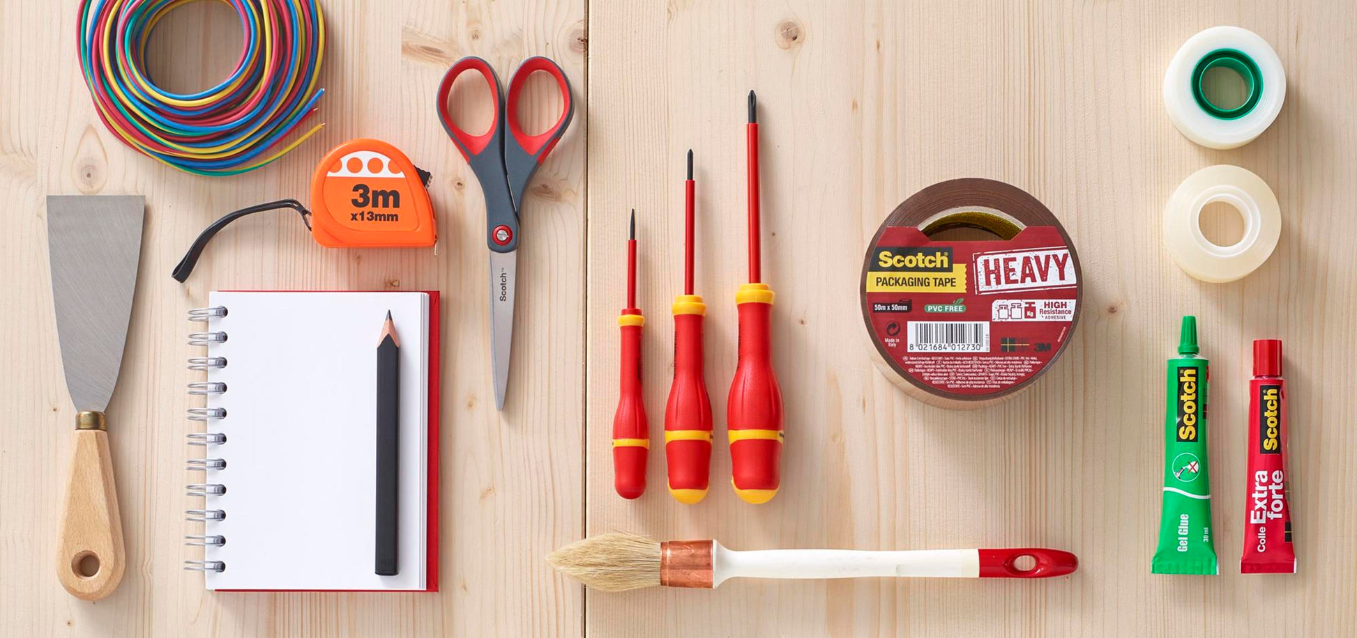 Eine Scotch™ Präzisionsschere rot der 3M Deutschland GmbH ist mit verschiedenen Werkzeugen und Schreibwaren wie farbigem Draht, Maßband, Schraubendreher, Klebeband, Spachtel, Spiralheft, Bleistift, Klebetube und Pinsel auf einer Holzfläche angeordnet.