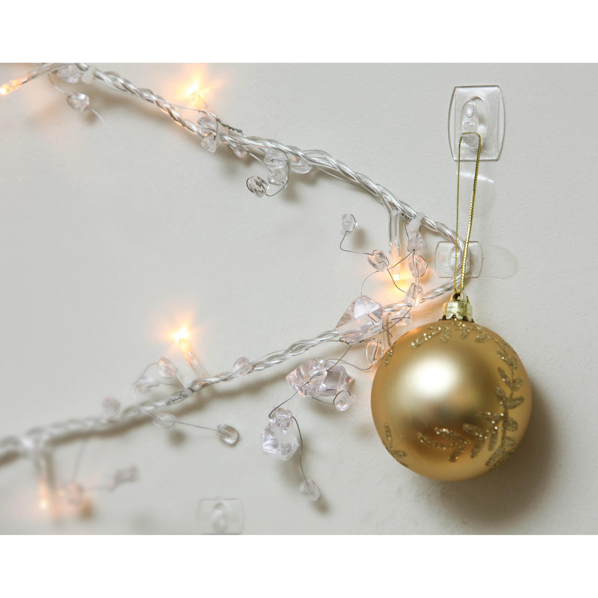 Ein goldener Weihnachtsschmuck hängt an einem Command™ Mini-Haken, transparent (3M Deutschland GmbH), der an einer weißen Wand neben weiß leuchtenden, mit Kunststoffkristallen verzierten Weihnachtslichtern angebracht ist.