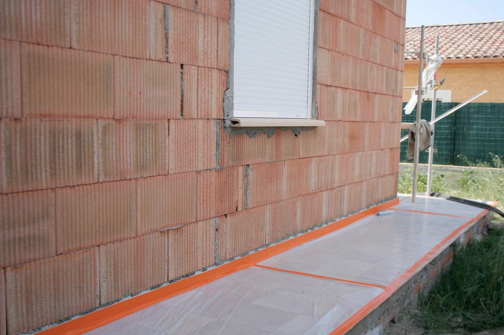 Eine gemauerte Außenwand mit einem geschlossenen weißen Fensterladen. Der Boden ist mit Kunststofffolien bedeckt, die mit Scotch® High Visibility Klebeband, orange, 25 m x 48 mm von 3M Deutschland GmbH gesichert sind, wahrscheinlich für Bau- oder Malerarbeiten.