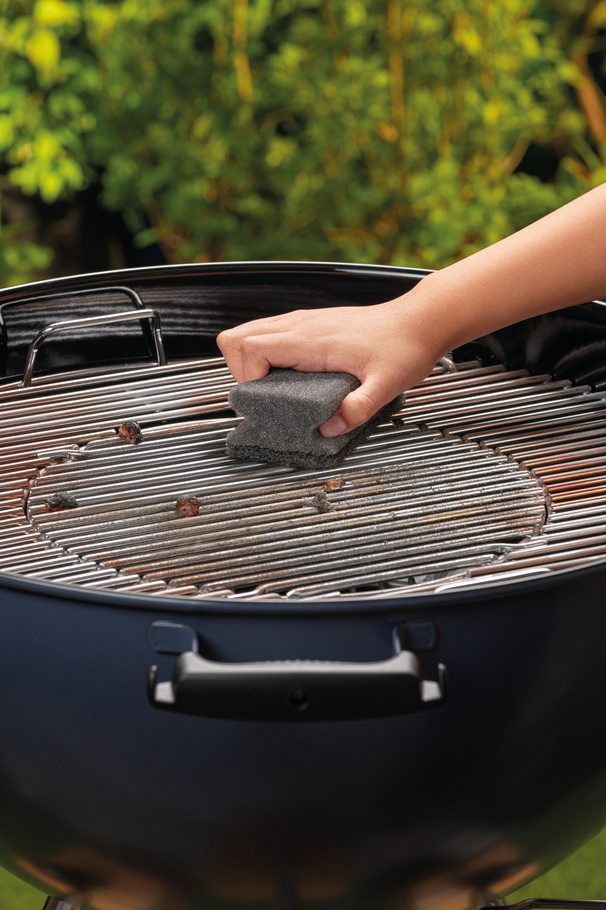 Eine Person schrubbt mit einem Scotch-Brite® Grill Griffschwamm von 3M Deutschland GmbH die Metallroste eines Holzkohlegrills im Freien, im Hintergrund ist eine Grünfläche zu sehen. Die Packung enthält 1 Stück.