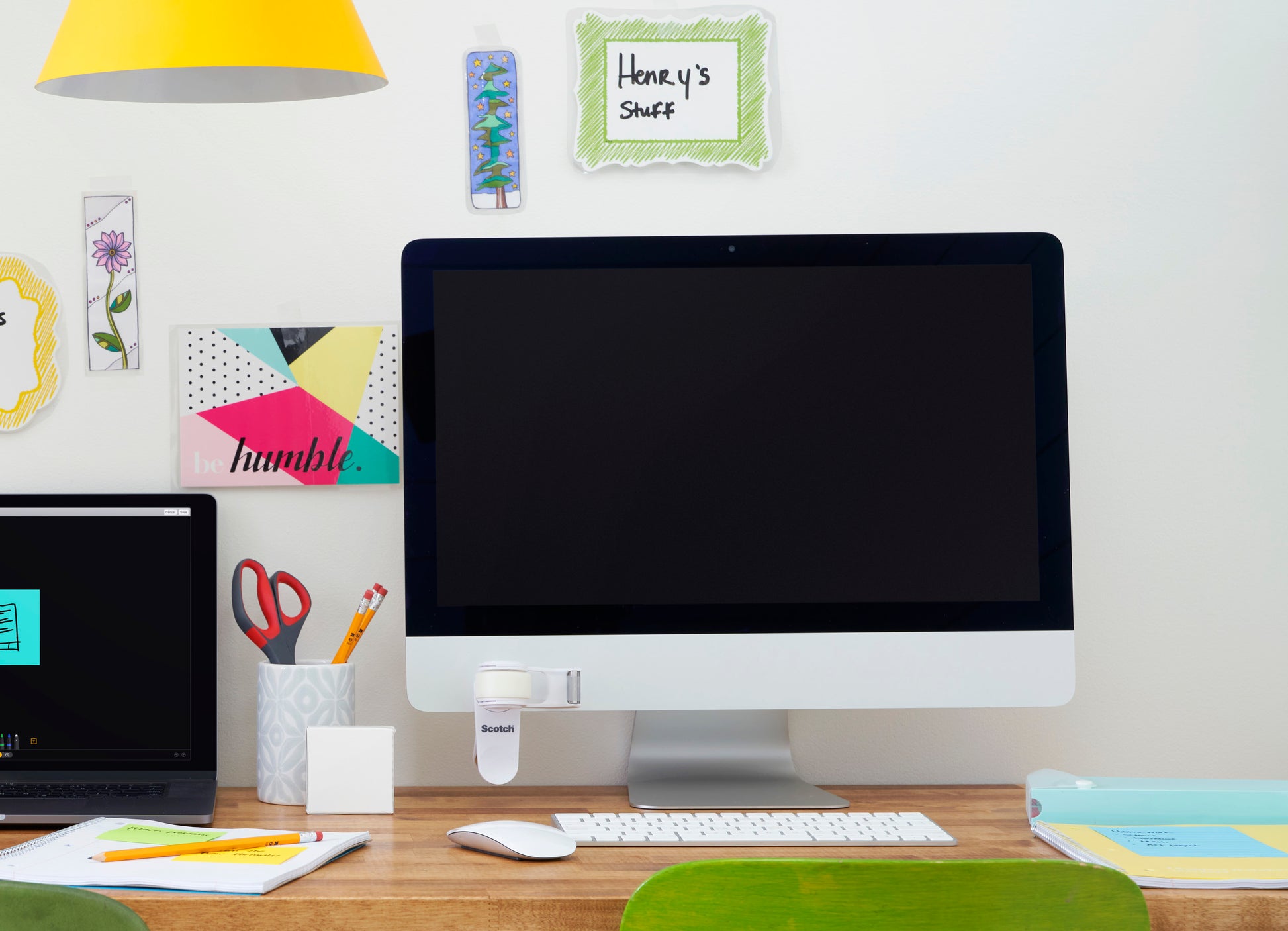 Ein aufgeräumter Schreibtisch mit einem großen Monitor, einem geschlossenen Laptop, einem Becher mit Stiften und Scheren, Büromaterial und dem 3M Scotch™ Clip & Twist Klebeband-Abroller C19 mit Magic™ Unsichtbarem Klebeband auf dem Display. An der Wand befinden sich dekorative Notizen wie "Henry's Stuff".
