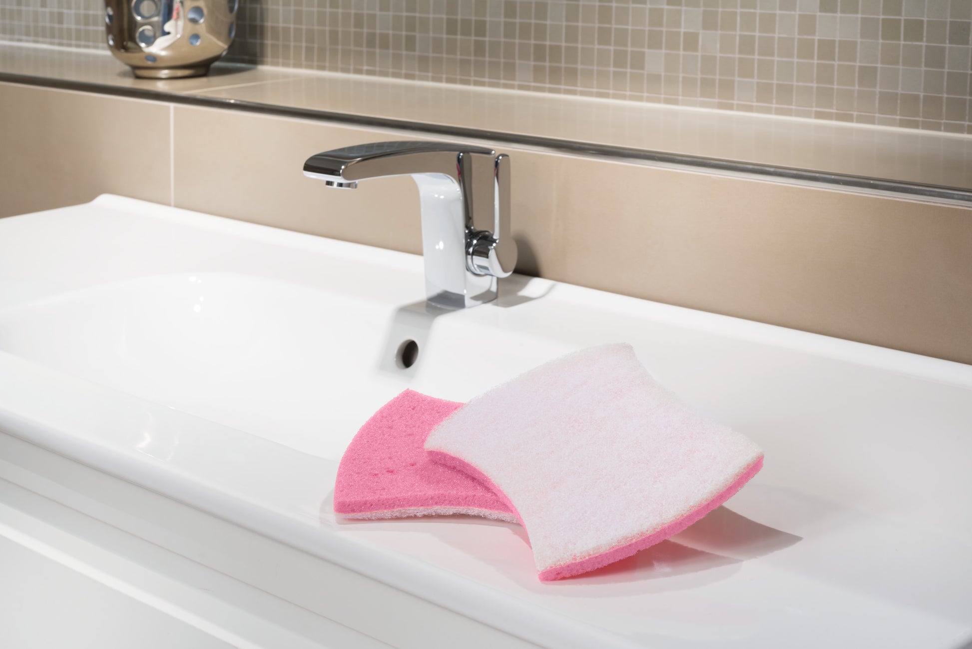 Eine moderne verchromte Armatur und ein weißes Waschbecken mit einem Scotch-Brite® Bath XXL Reinigungsschwamm für das Bad (1 Stück) von 3M Deutschland GmbH auf der Arbeitsplatte vor beigen Fliesen.
