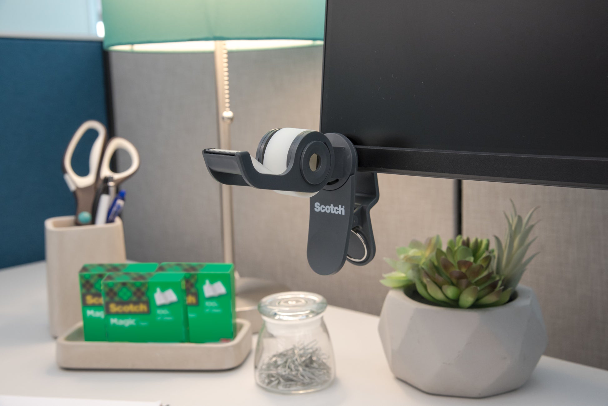 Auf dem Schreibtisch: ein 3M Scotch™ Clip & Twist-Klebebandabroller mit Scotch® Magic™ Invisible Tape (19 mm × 8,89 m), eine Lampe, eine Schere in einem Halter, zwei zusätzliche Rollen Scotch®-Klebeband, ein Glas mit Stecknadeln und eine kleine Sukkulentenpflanze im Topf.